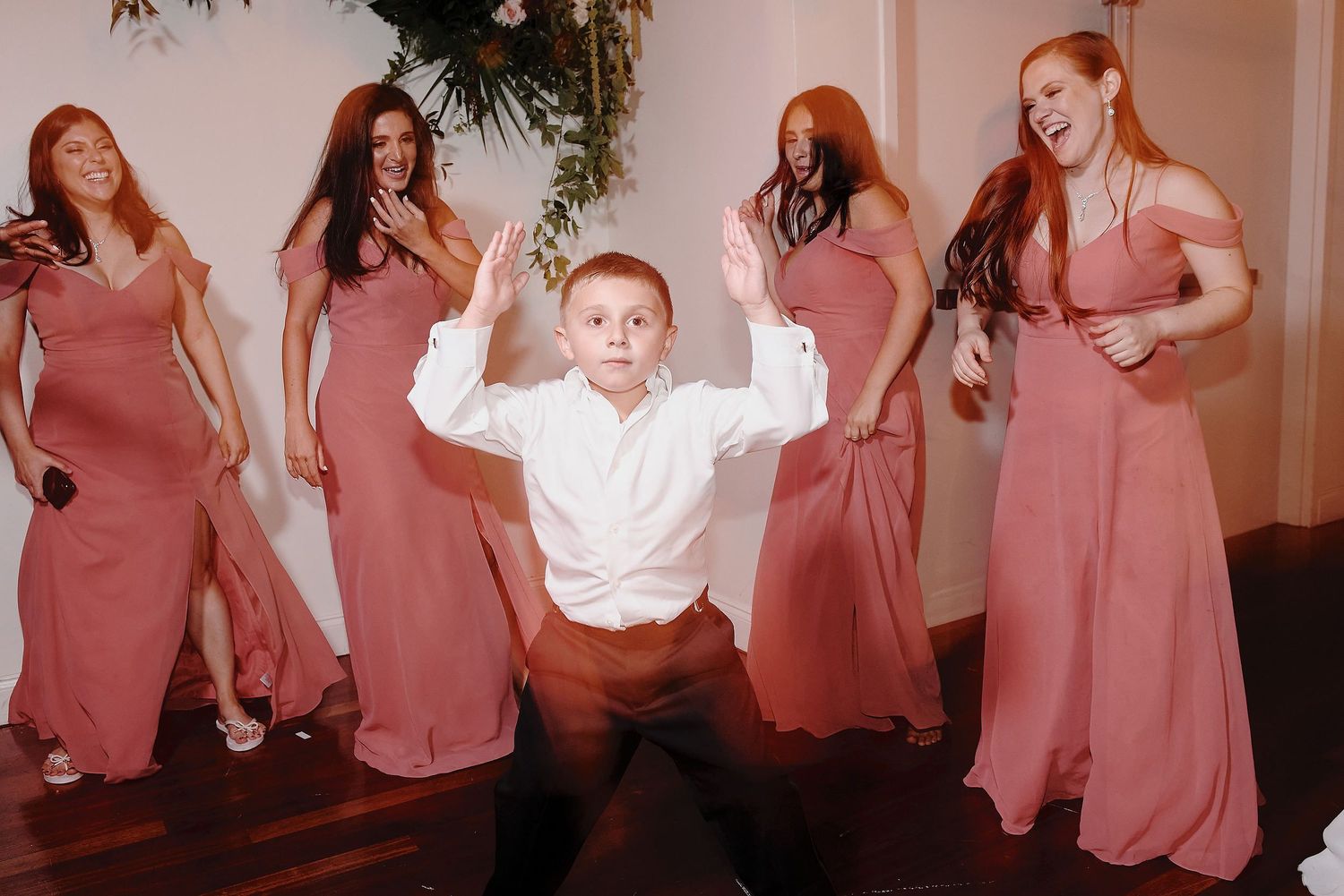 Young boy in white shirt dances energetically surrounded by bridesmaids in dusty rose dresses at a wedding reception.
