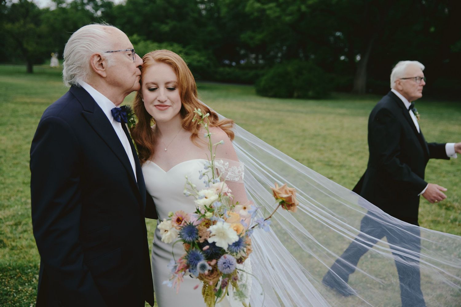 A wedding scene outdoors in a green field with flowing veil and floral bouquet.