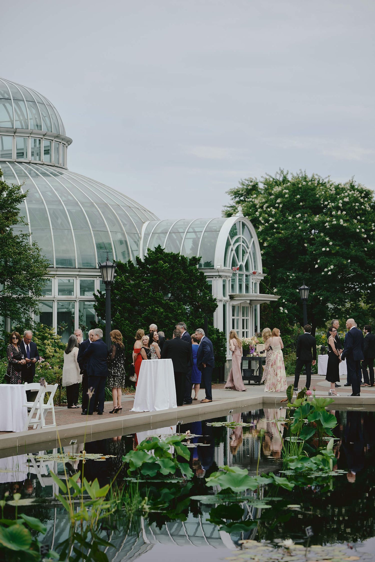Wedding guests gather for an elegant reception by a reflecting pool outside a glass conservatory dome at a botanical garden.
