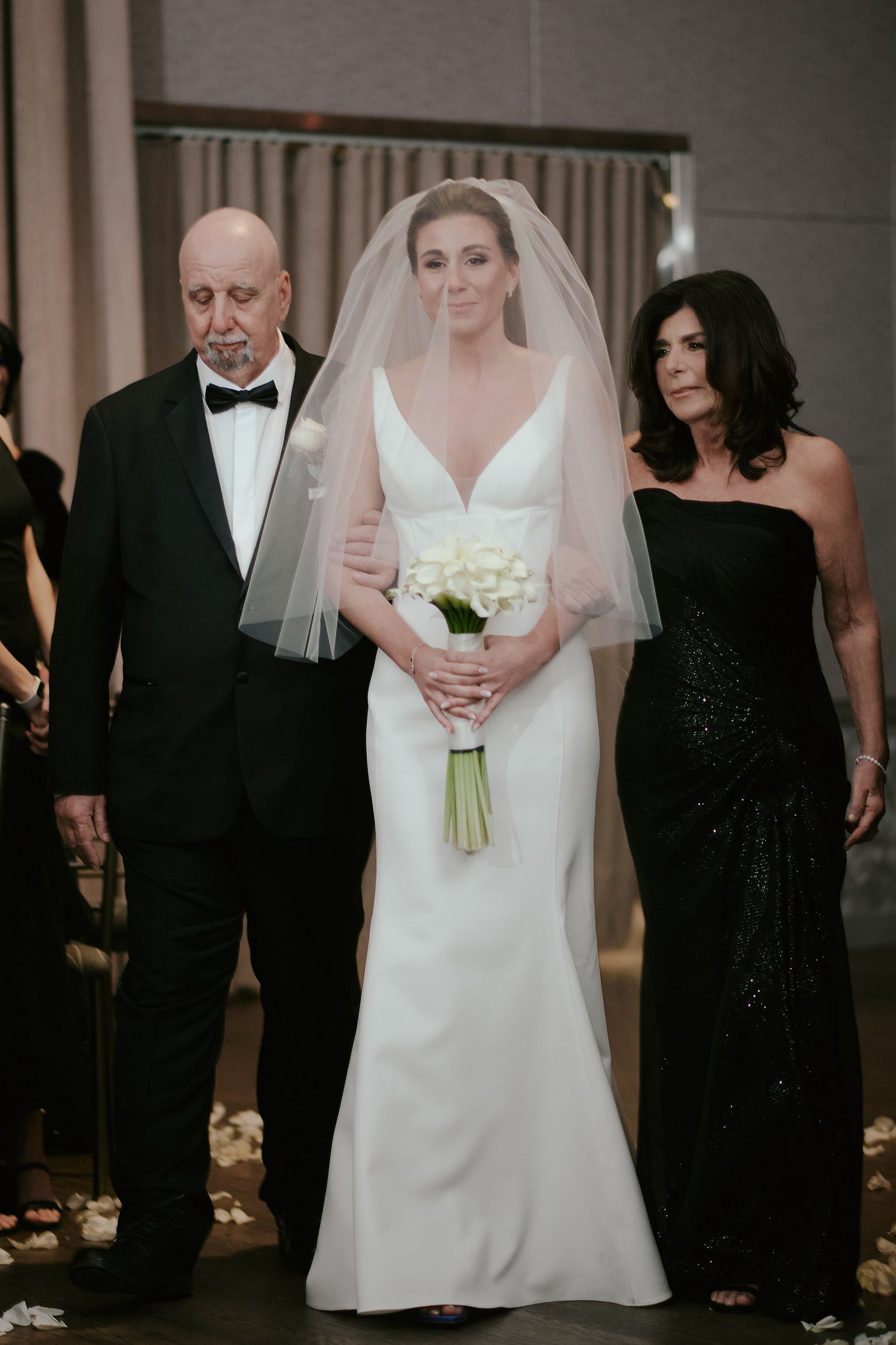 A bride in a classic white gown walks down the aisle with her bouquet, accompanied by parents in formal black attire.