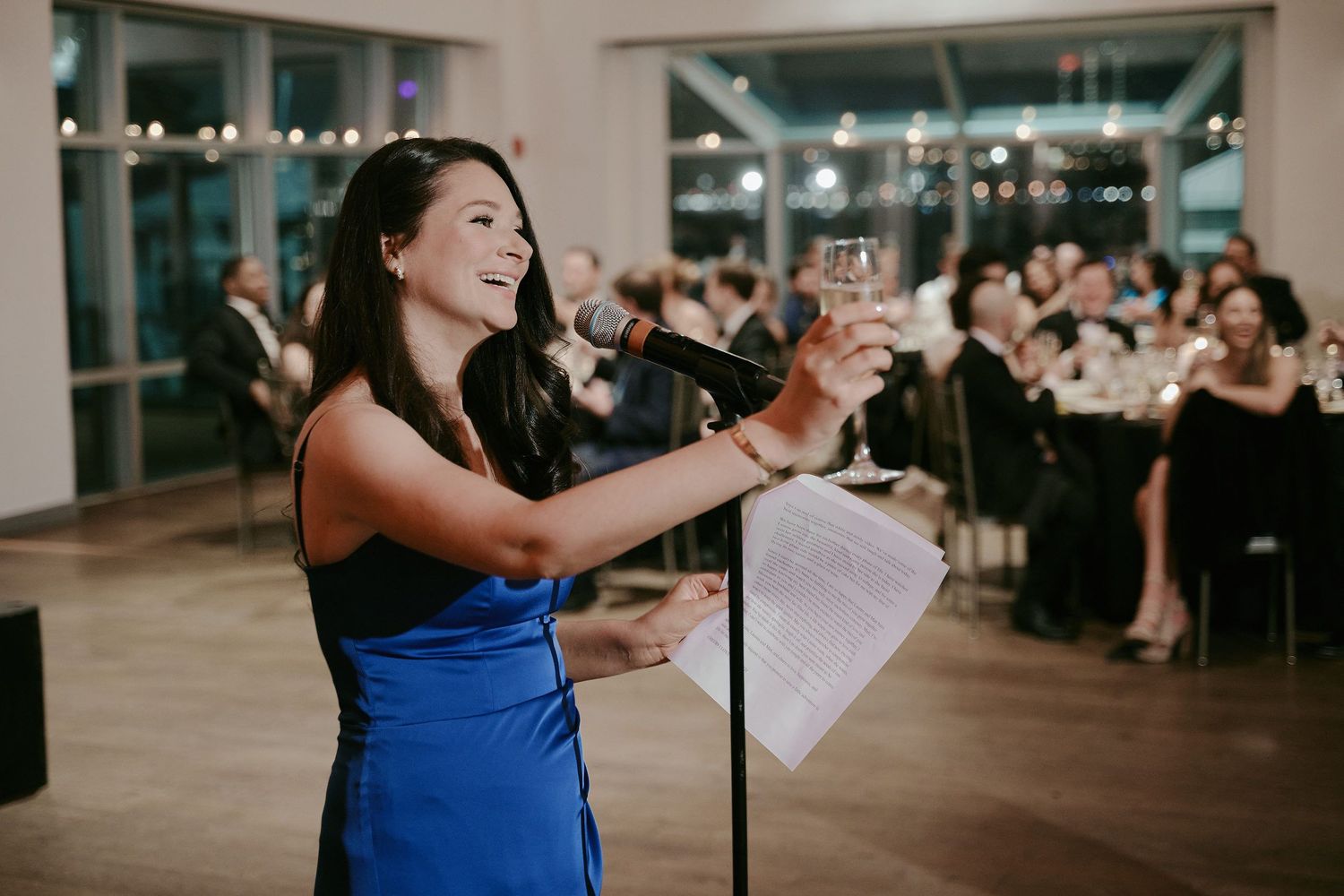 A performer in a royal blue dress sings from sheet music at a formal event with guests seated in the background.
