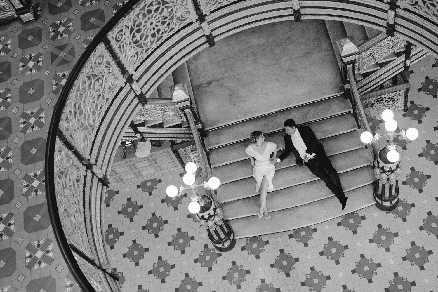 Aerial black and white photo of a couple dancing on grand staircase with ornate chandelier and tiled floor.