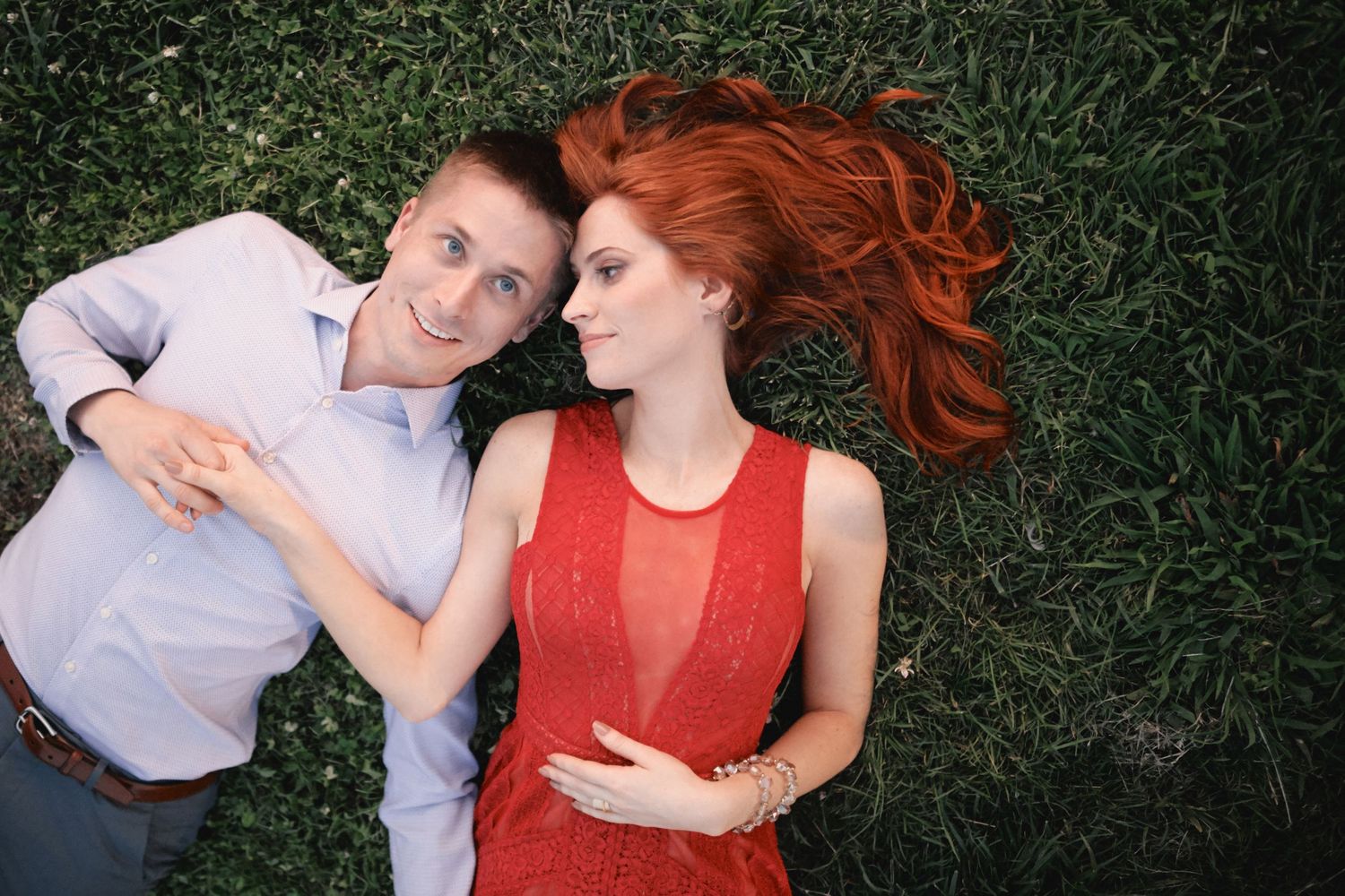 A couple lies peacefully together on green grass, with one wearing white and the other in a vibrant red sleeveless dress.