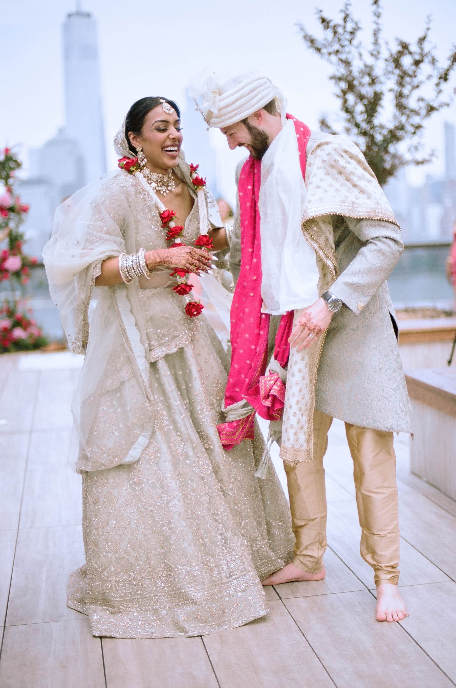 A wedding couple in traditional Indian attire sharing a joyful moment on a rooftop with city skyline in background.