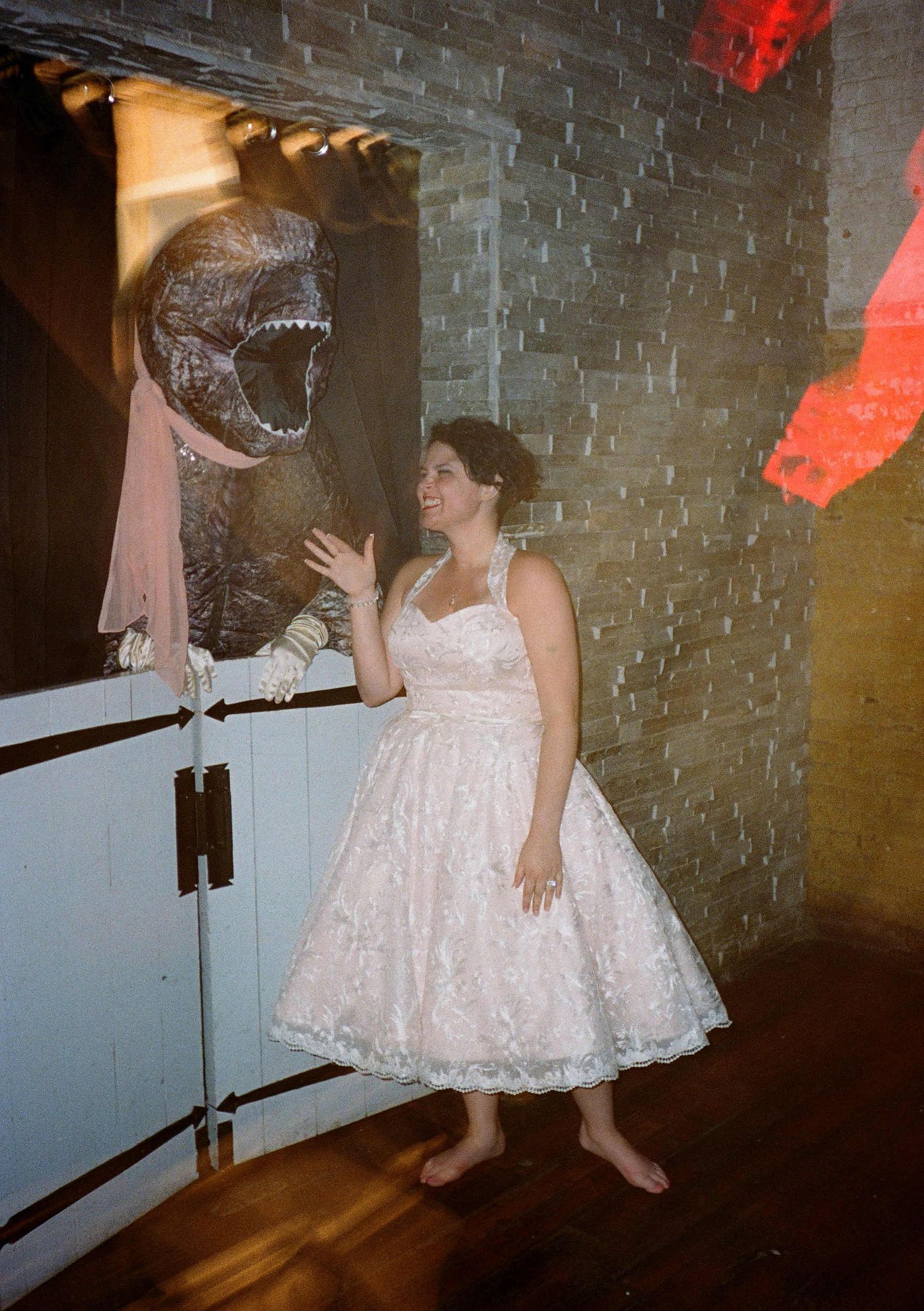 A person in a white lace tea-length dress poses with a large silver balloon in a brick-walled room at a party.