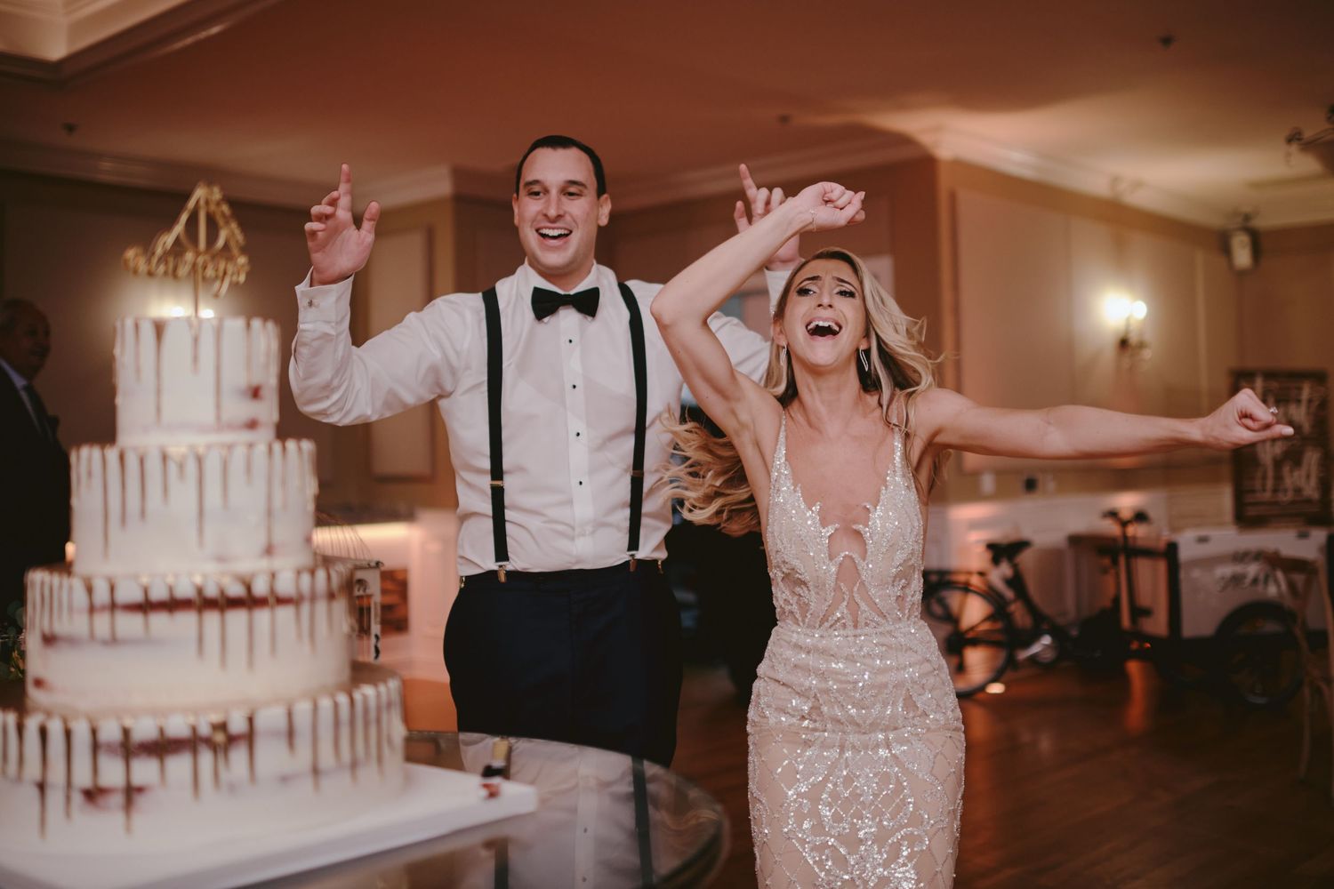 Newlyweds celebrating with joyful arms raised next to their elegant white tiered wedding cake at reception.
