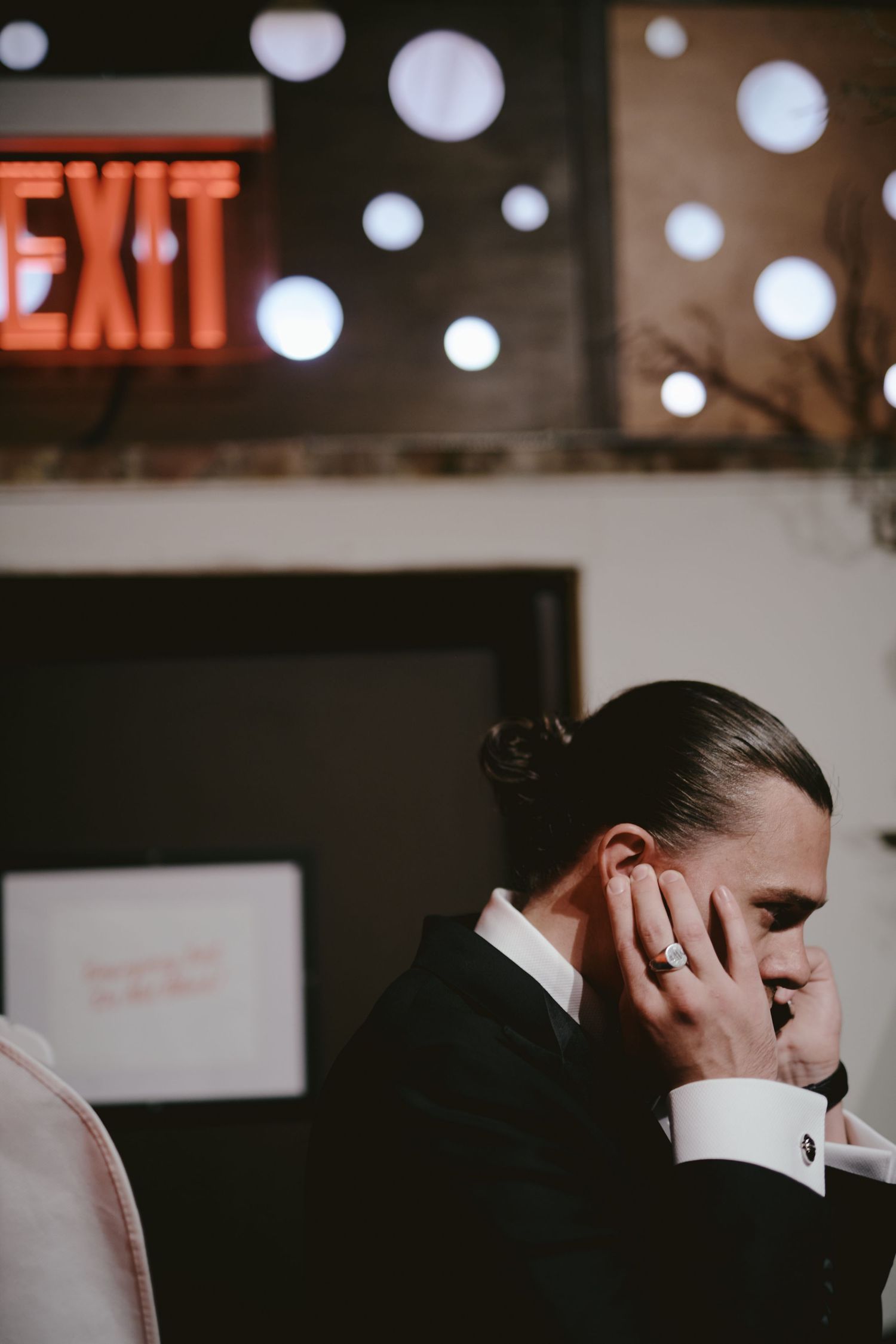 A person in formal attire sits with a downturned gaze beneath a glowing red exit sign and decorative spotlights.