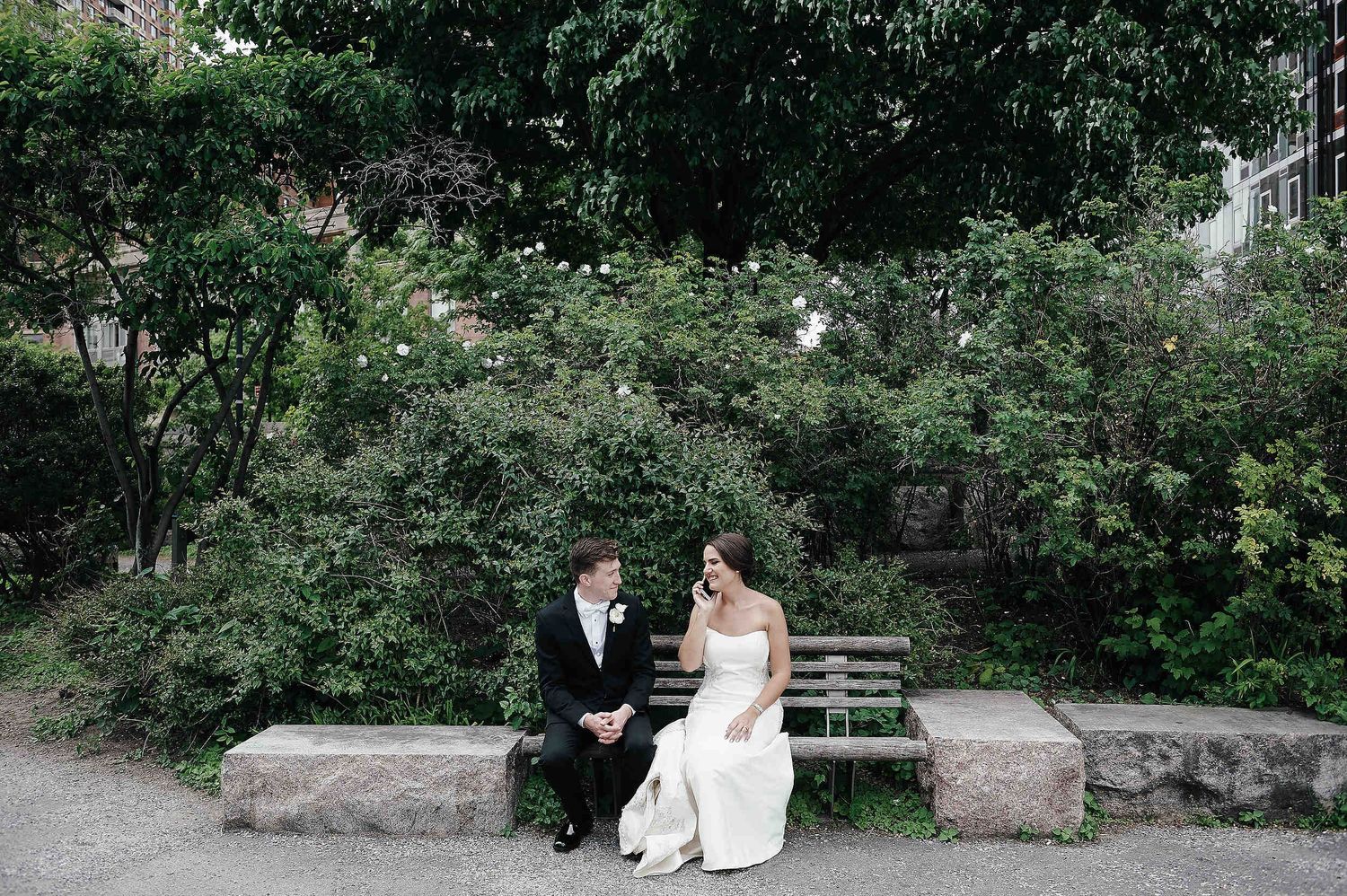 Two people in formal attire sit together on a garden bench surrounded by lush greenery and flowers.