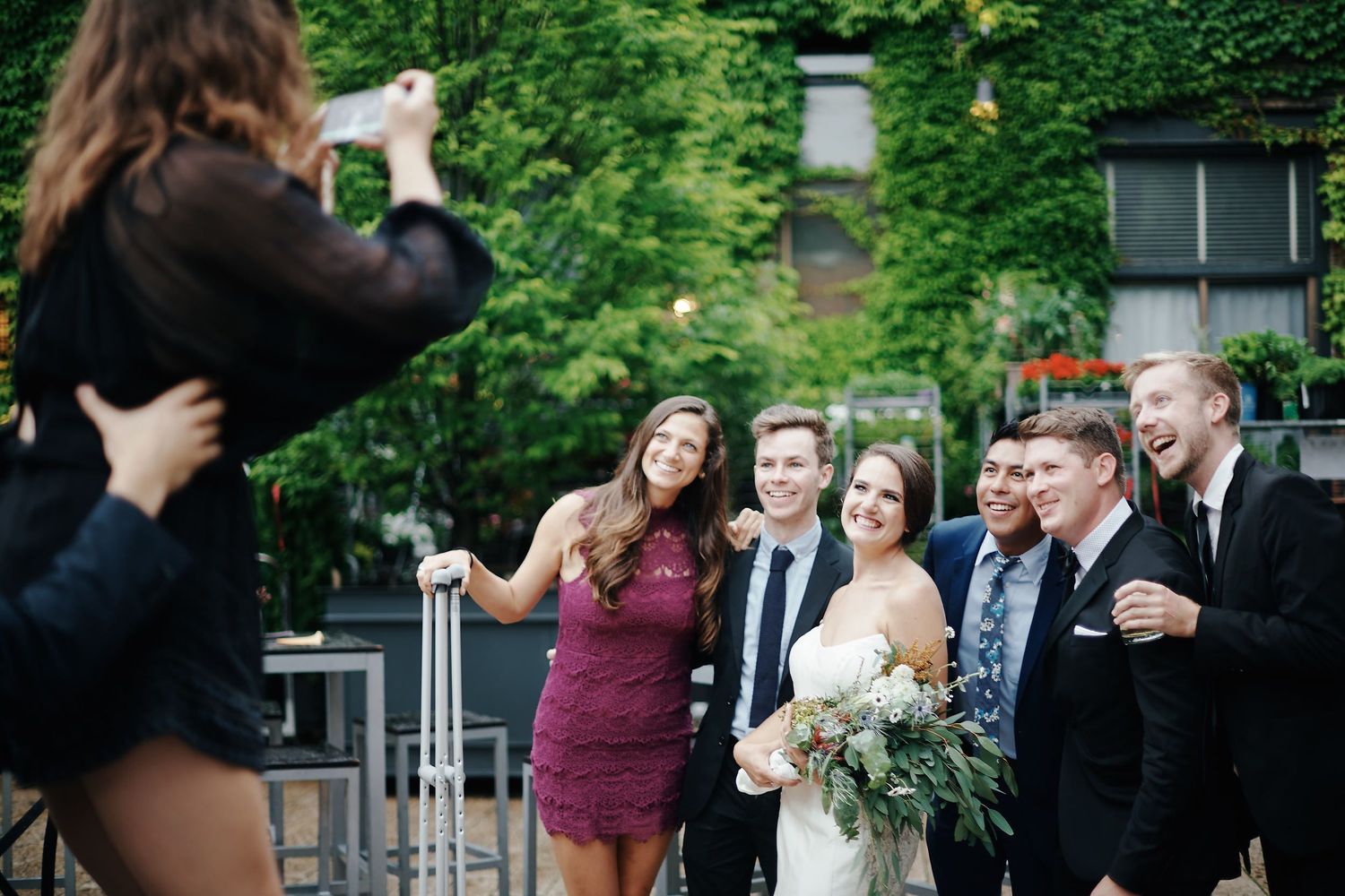 A photographer captures a wedding party posing outdoors with a lush green garden backdrop.