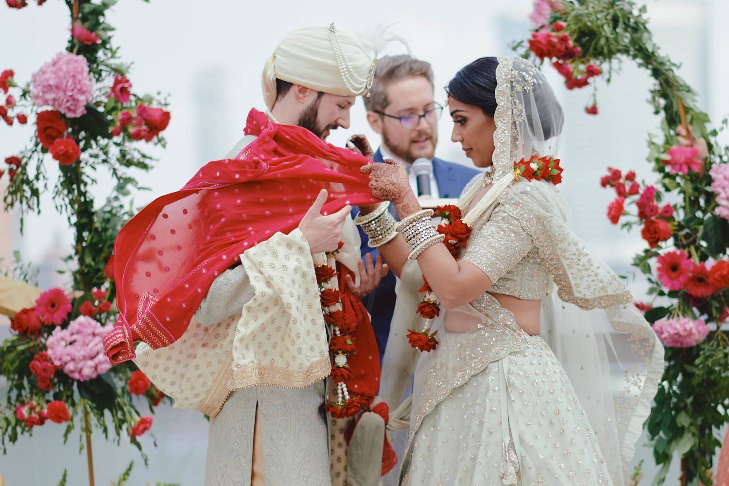 A traditional wedding ceremony under a floral arch with pink and red roses, featuring participants in ornate white and red attire.