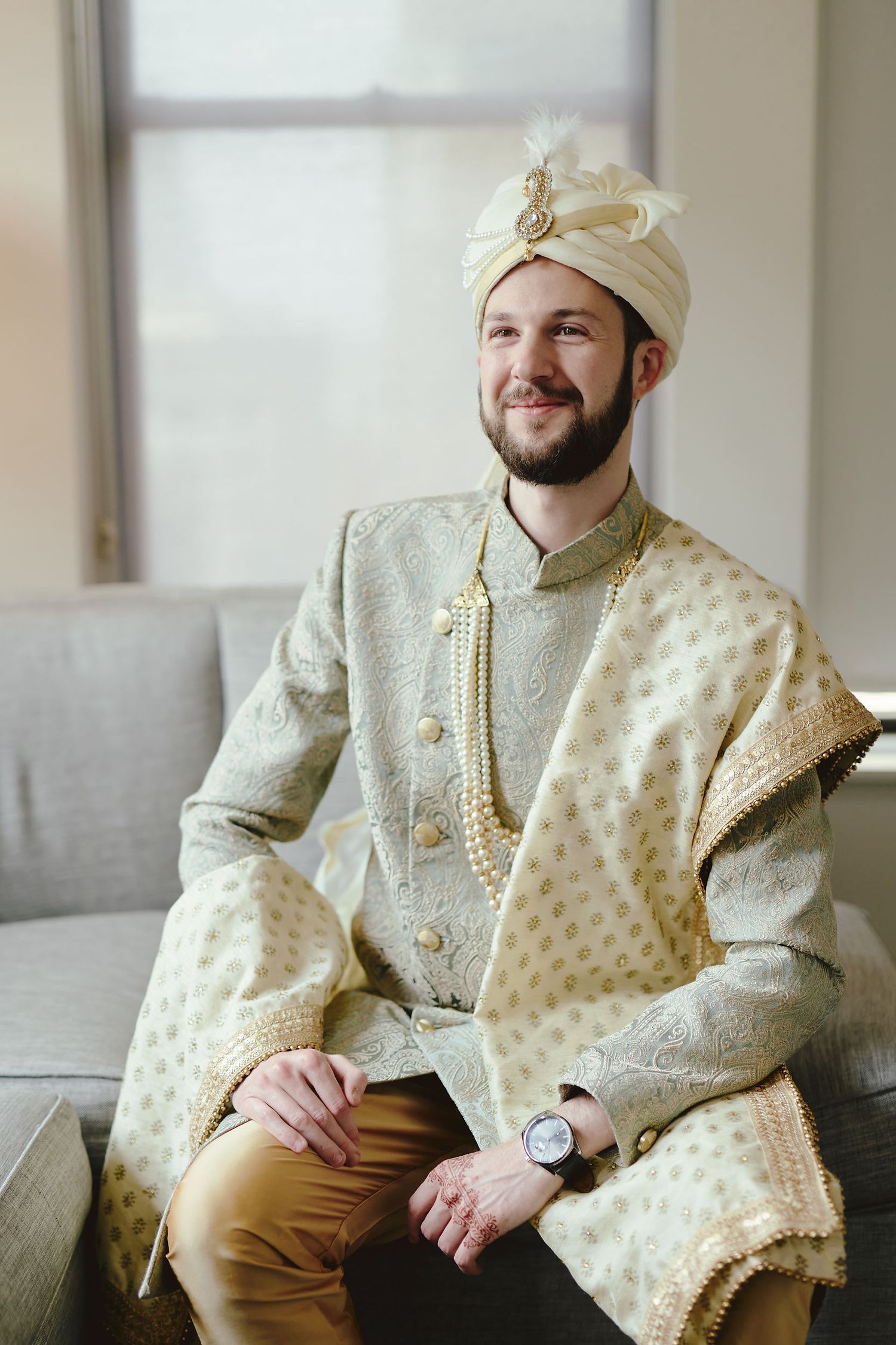 Groom wearing traditional Indian sherwani in cream and gold with matching turban sits elegantly on couch.