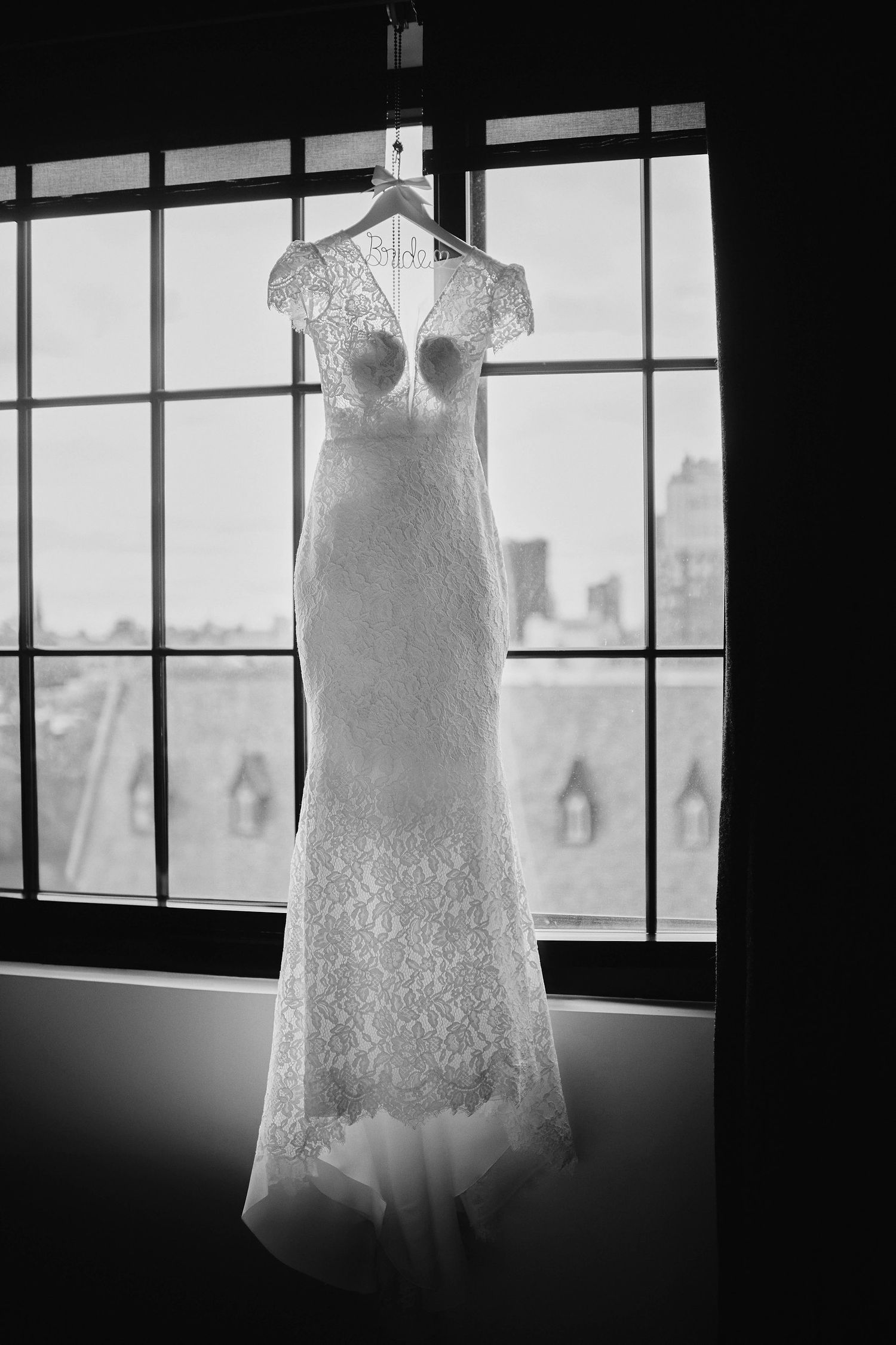 A vintage lace wedding dress with a fitted silhouette hangs in front of a window in black and white.