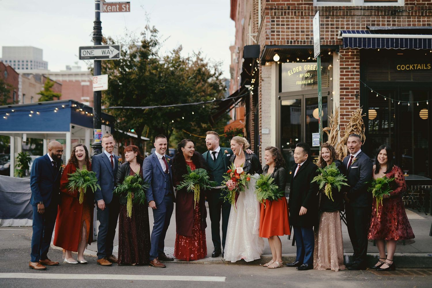 Wedding party in formal attire stands together on city street corner holding green bouquets at evening celebration.