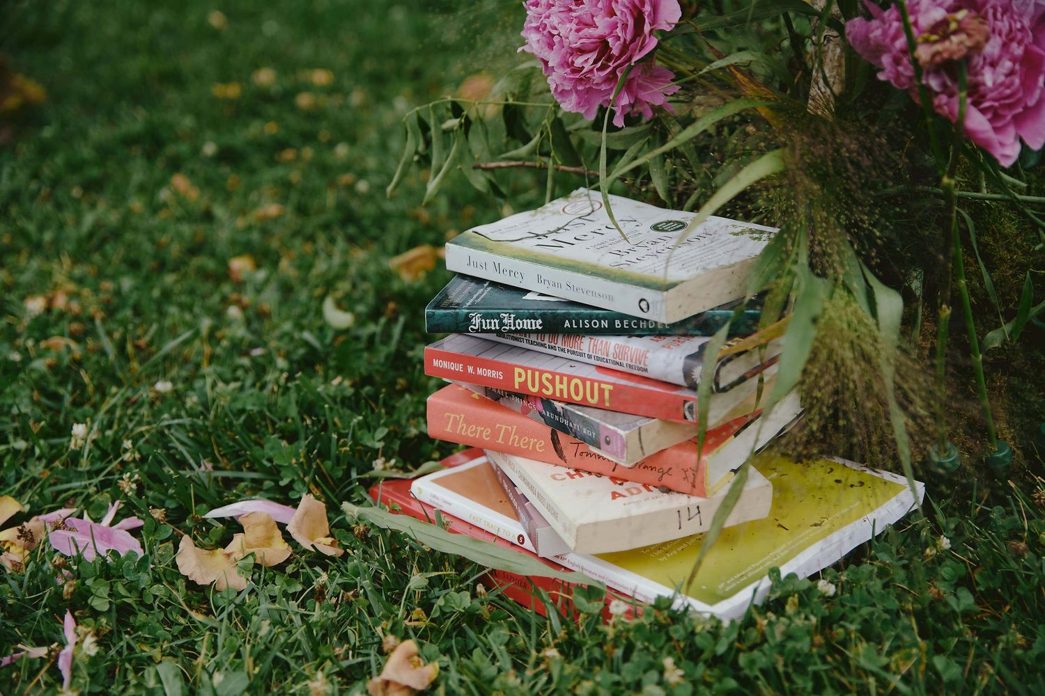 Stack of books laying on green grass next to blooming pink peonies in a garden setting.