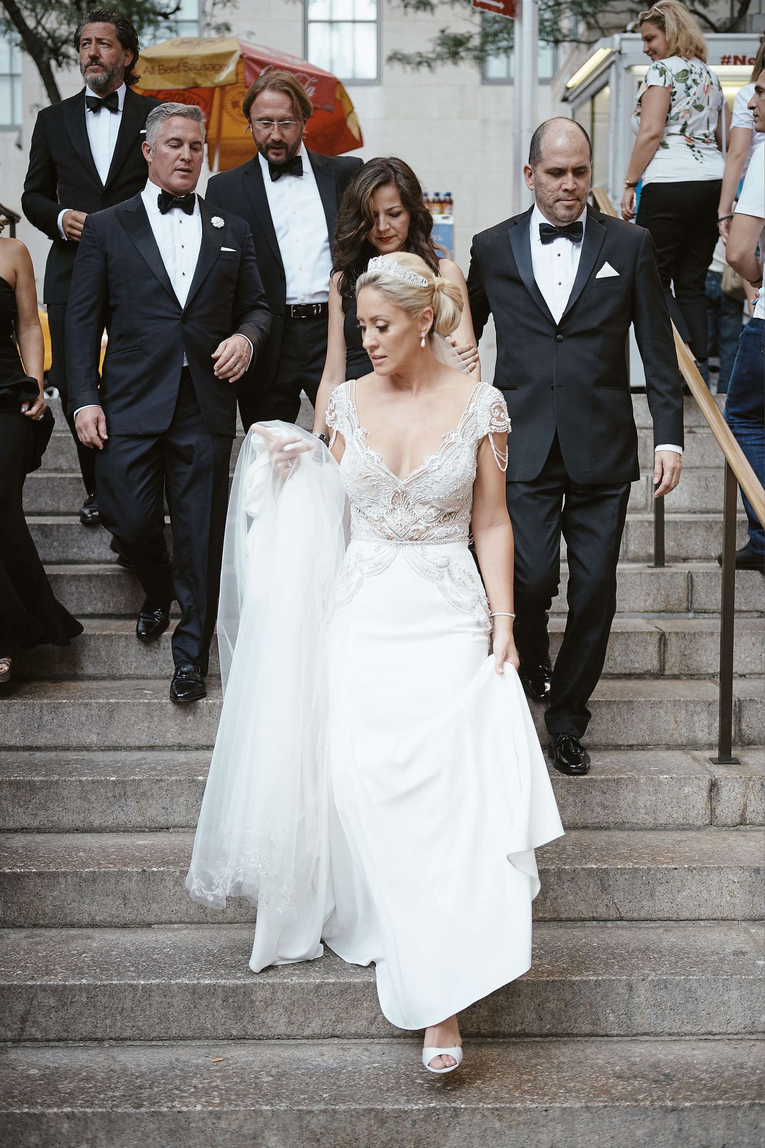 Bride in elegant lace V-neck wedding dress descends stone steps accompanied by formally dressed guests at outdoor venue.