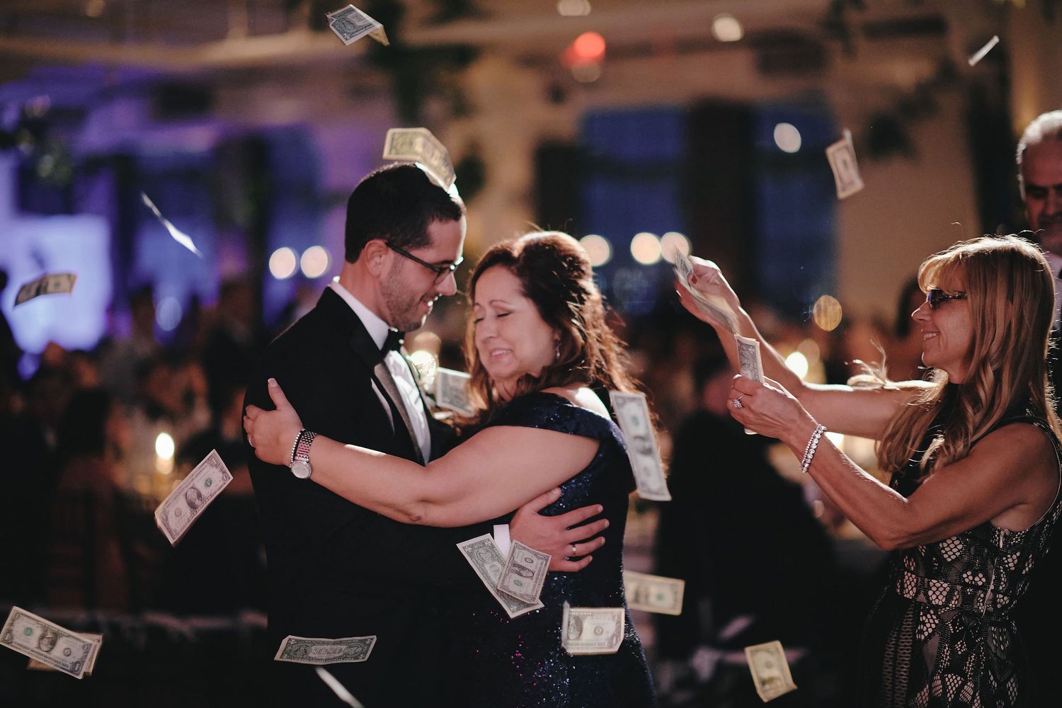 Newlyweds share their first dance while guests celebrate by tossing money in a dimly lit reception venue.