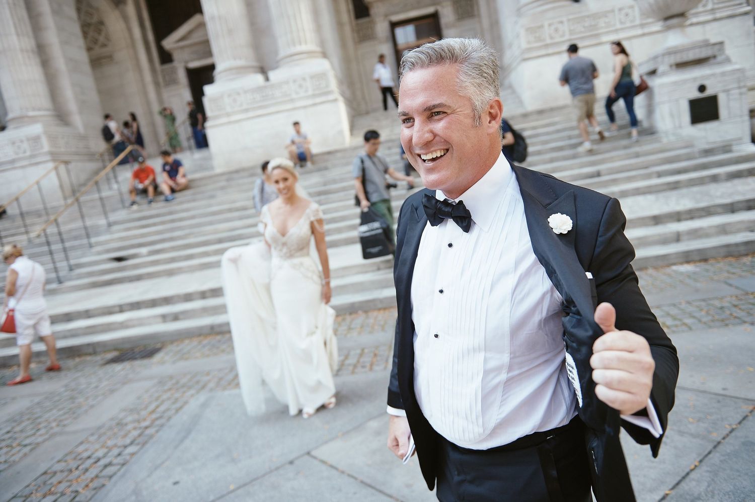 A happy groom in a tuxedo gives a thumbs up while walking down stone steps at a grand building entrance.