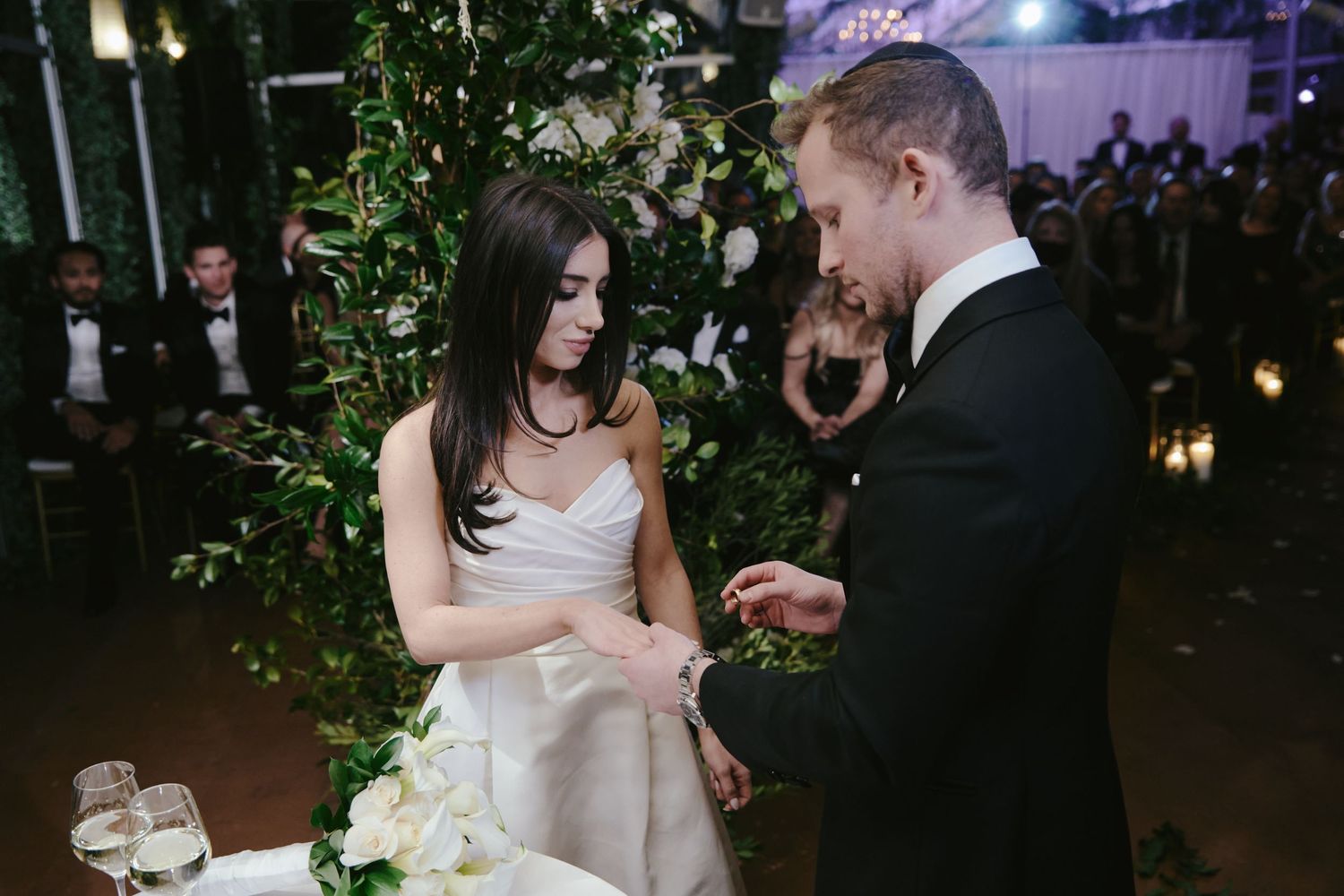 A romantic wedding moment captured during the ring ceremony amidst greenery and atmospheric lighting.