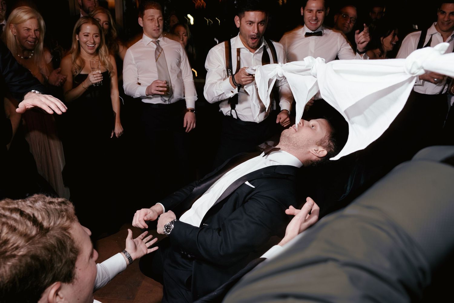 Wedding reception guests lift and carry someone in formal attire across the dance floor during celebration.