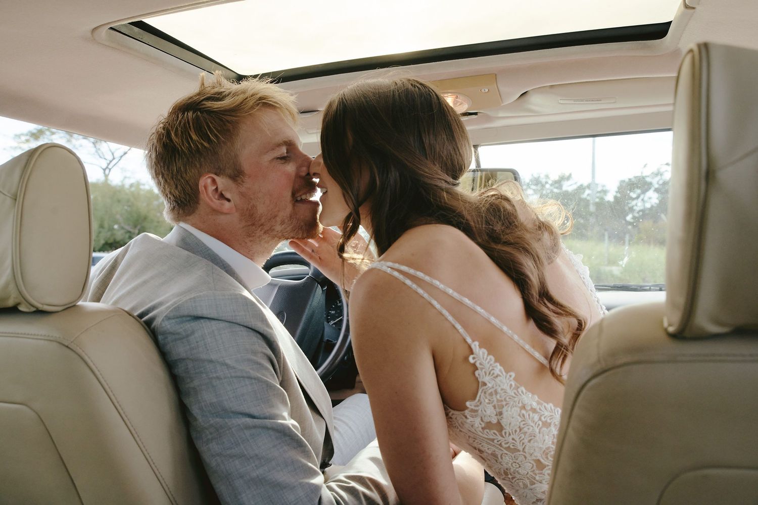 A couple shares a joyful moment together in the backseat of a car, with the bride wearing a white lace dress.