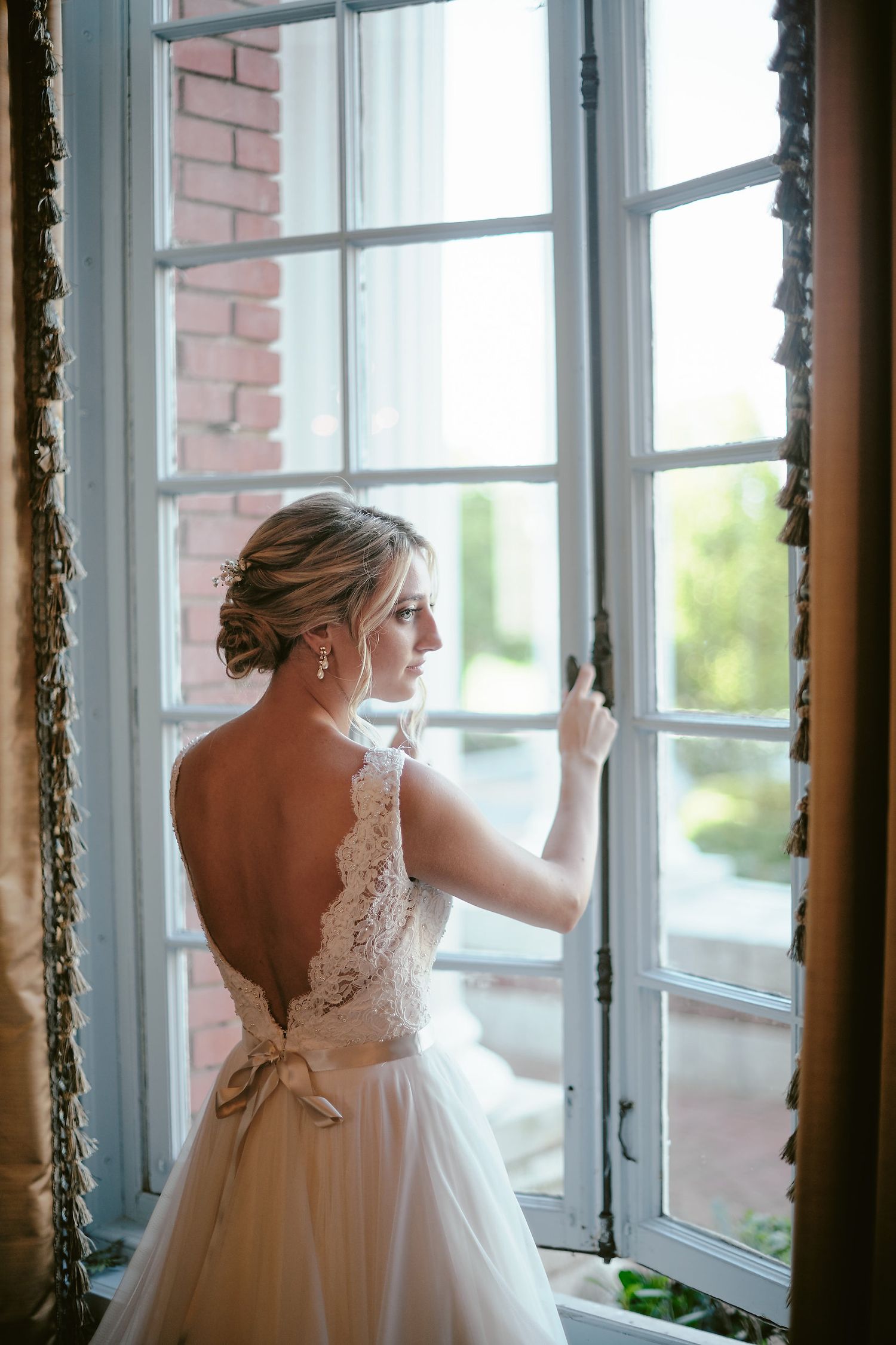 A bride in a backless lace wedding gown with a bow sash gazes through a vintage window with natural light streaming in.