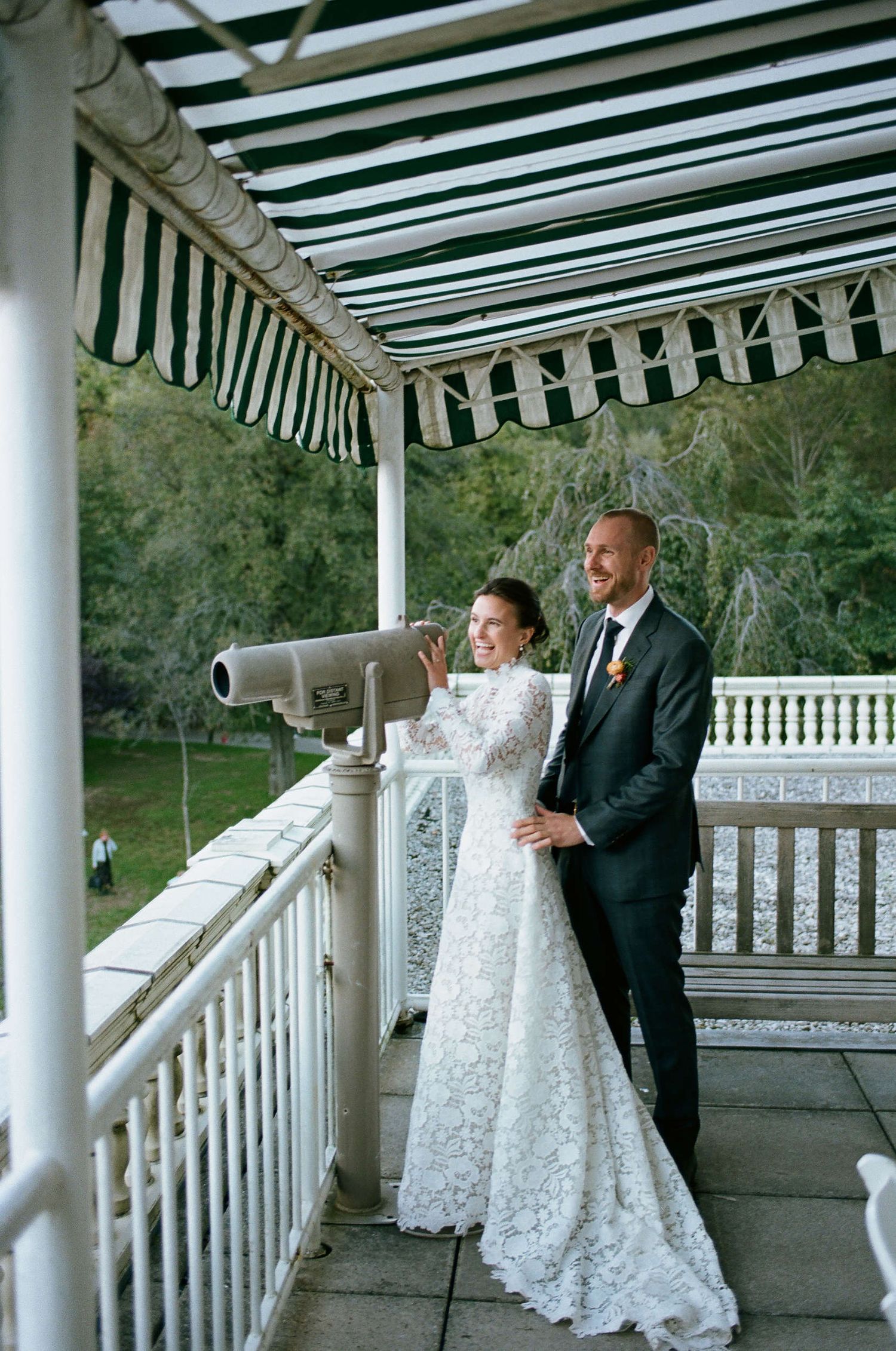 A couple stands on a white-columned porch with striped awning as they look through a mounted viewing telescope together.