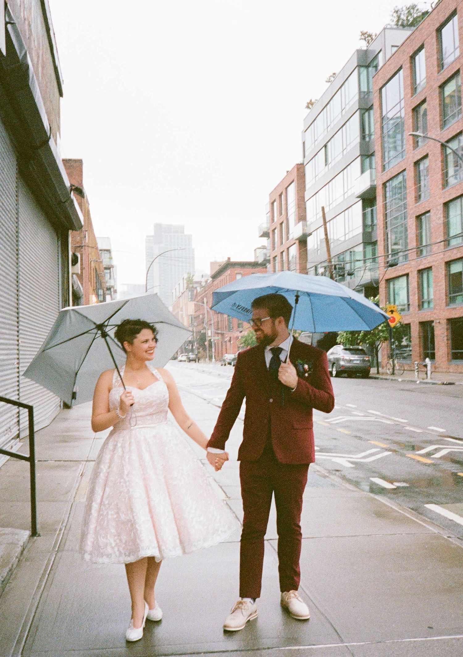 A couple in vintage wedding attire walk down a city street holding umbrellas on a rainy day.