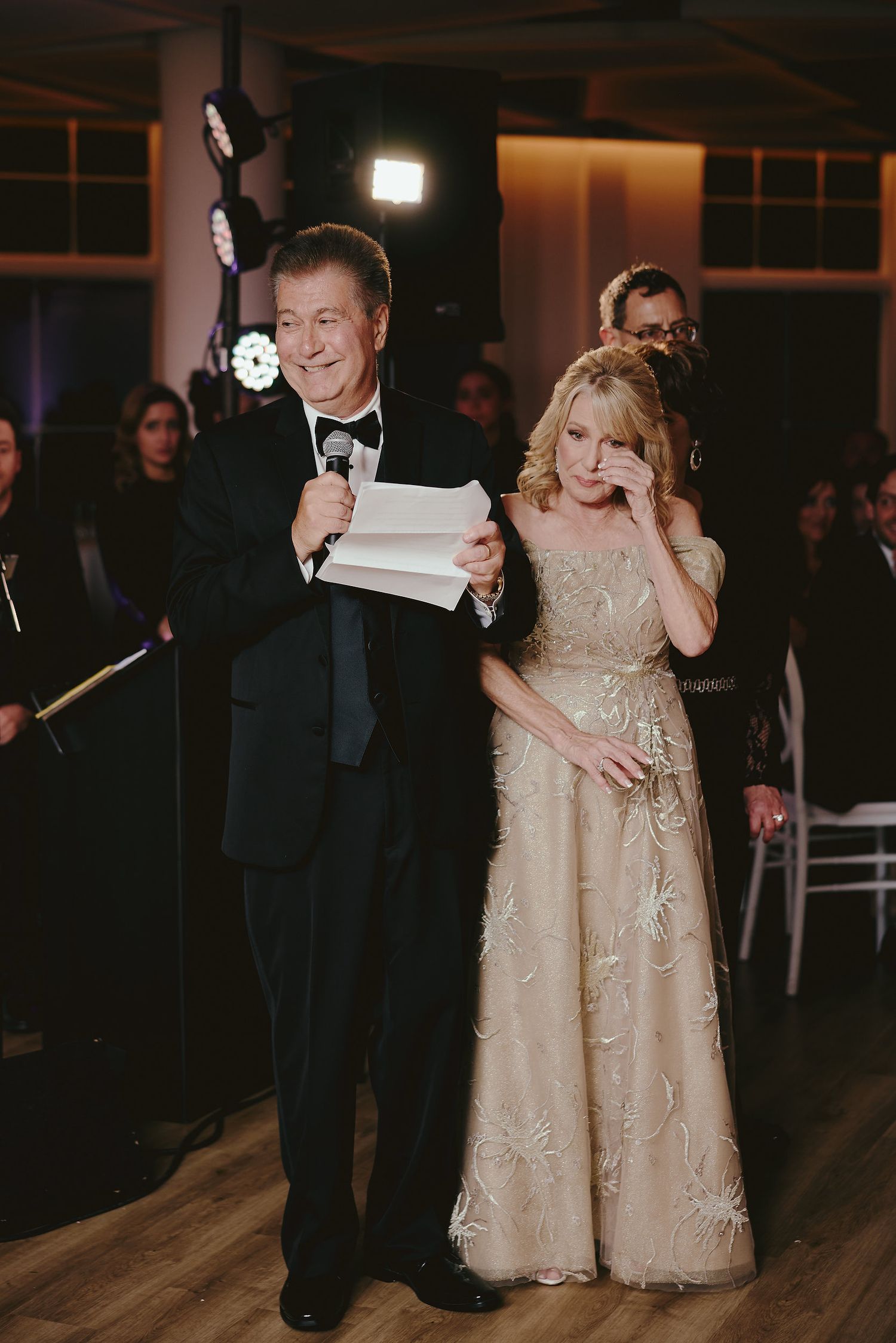 Couple at a formal event with the man in a tuxedo holding papers and woman in a champagne-colored gown.
