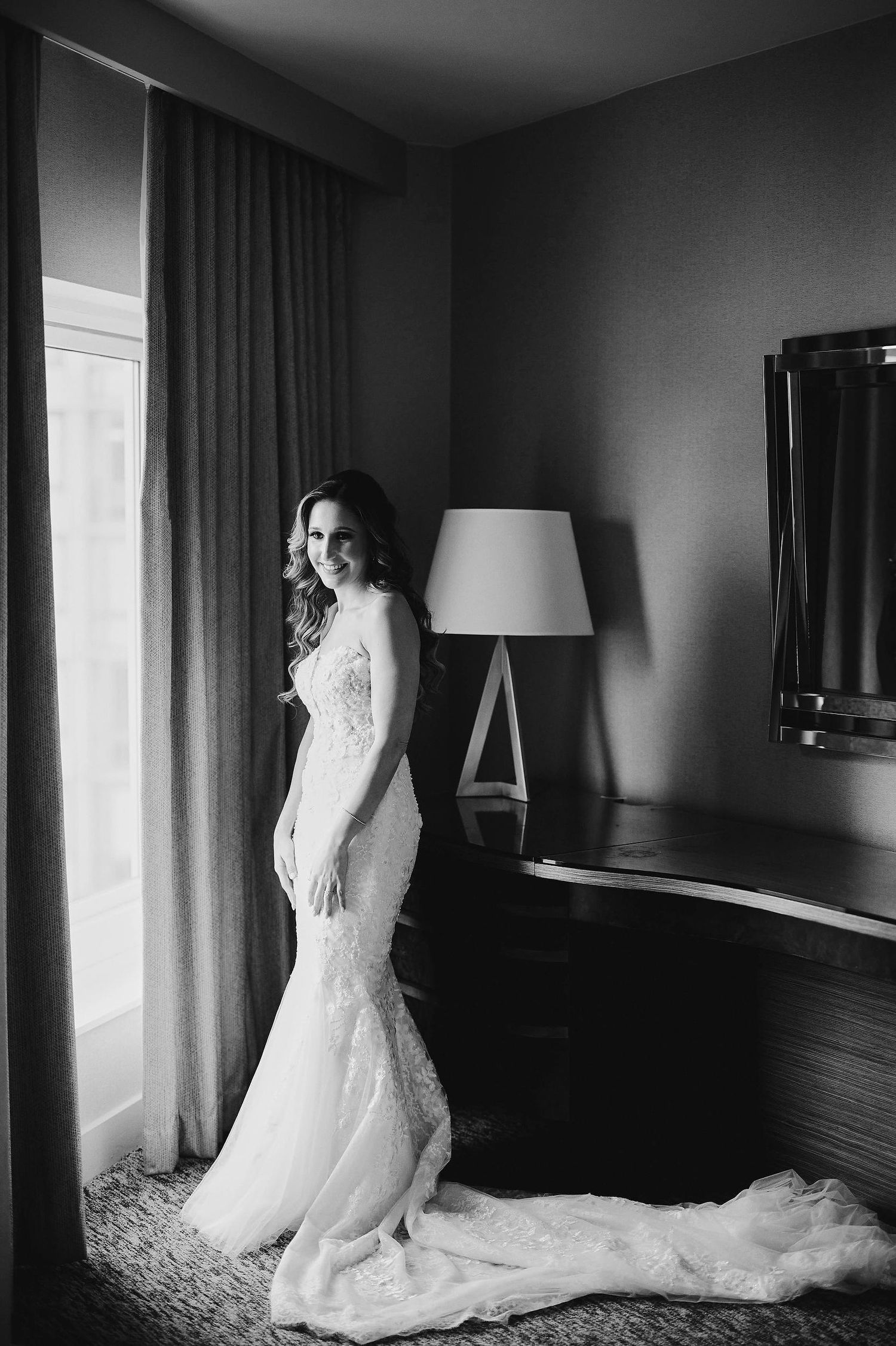 An elegant wedding dress with a fitted silhouette and long train photographed in a hotel room setting.
