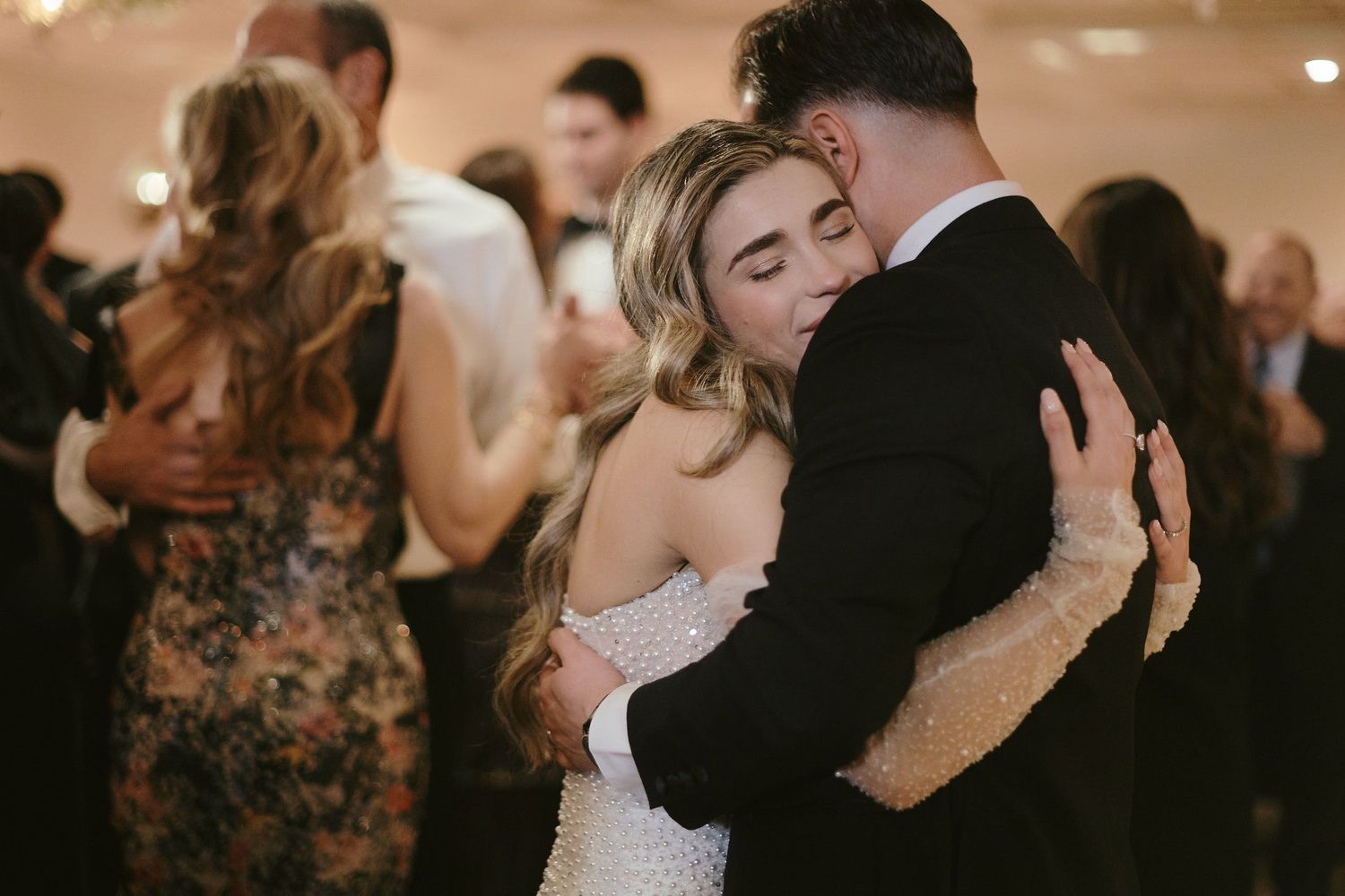 Two people slow dance together at a dimly lit wedding reception while other couples dance in the background.