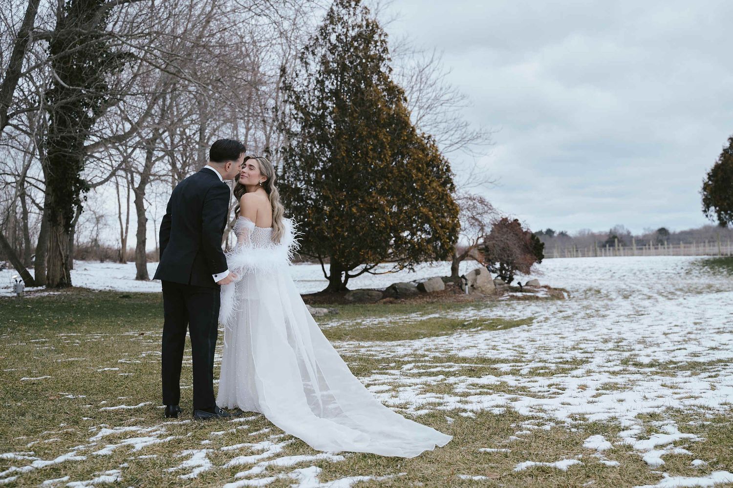 A couple in wedding attire stands together on snowy ground with trees and a wintry landscape in the background.