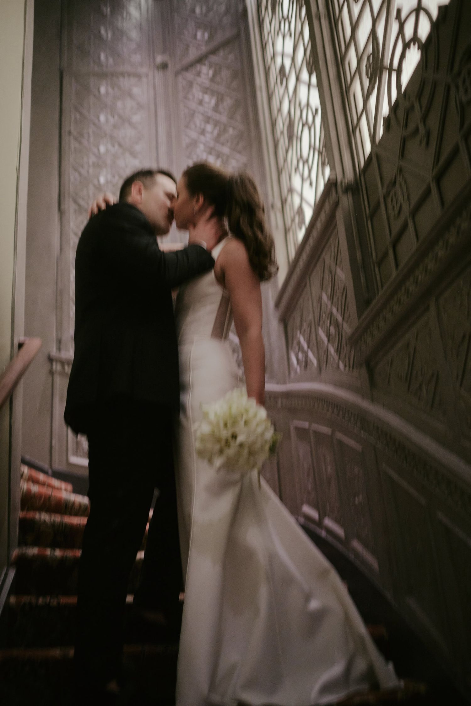 Romantic wedding couple embraces on elegant staircase with ornate detailing and white bouquet.
