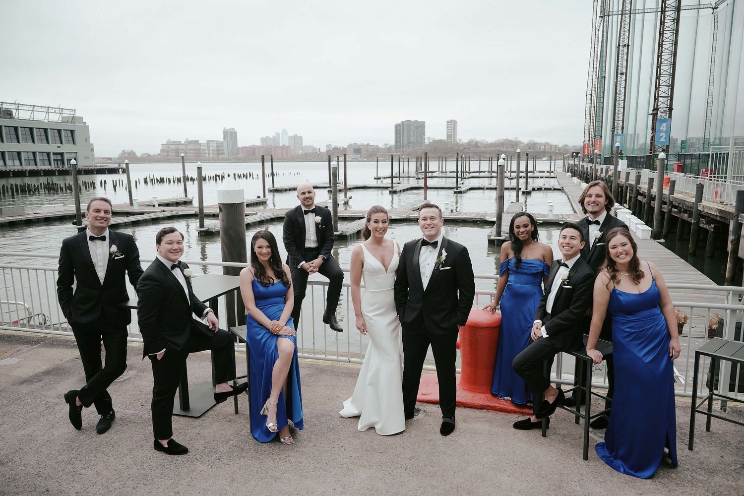 A wedding party poses together on a waterfront deck with city skyline and harbor in the background.