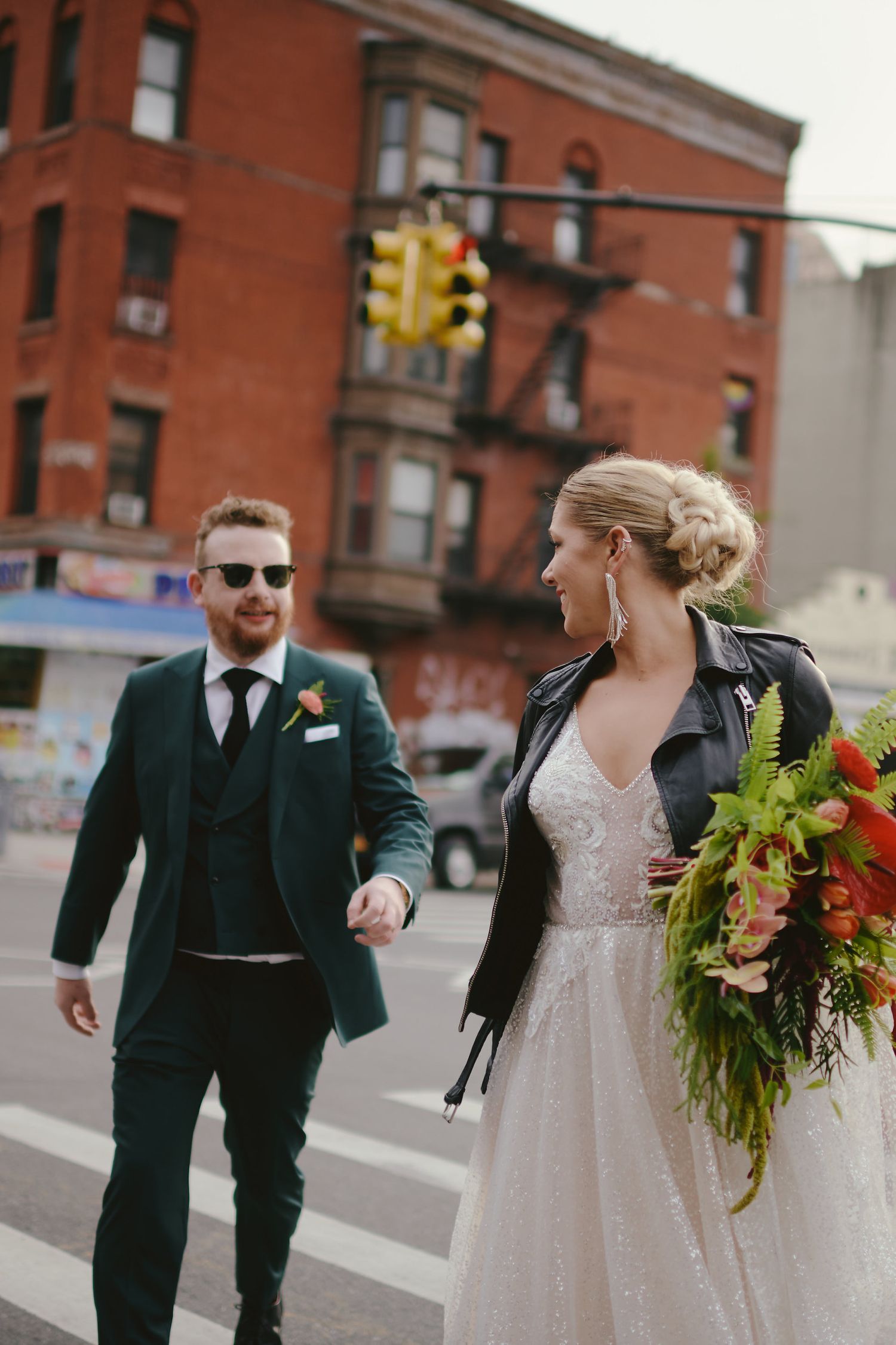 A couple in wedding attire walks across an urban street intersection with a brick building and traffic light in background.