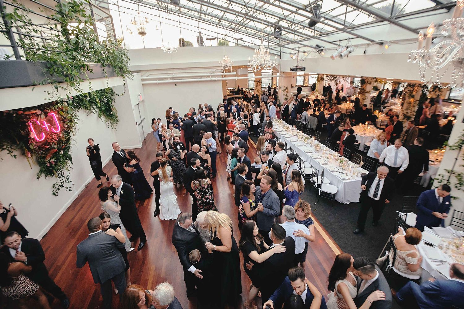 A large crowd of wedding guests mingle and celebrate in an elegant indoor venue with a glass ceiling and greenery.