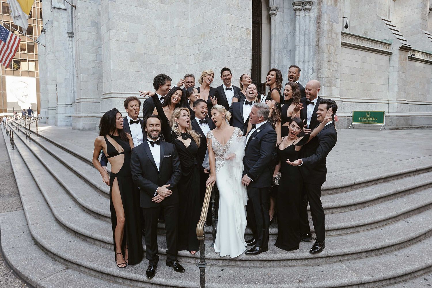 Wedding party wearing formal black and white attire poses on grand stone steps of a building.