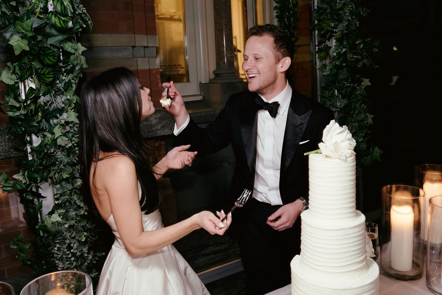 A couple shares a playful cake-tasting moment at their wedding reception next to a white tiered cake surrounded by candles.