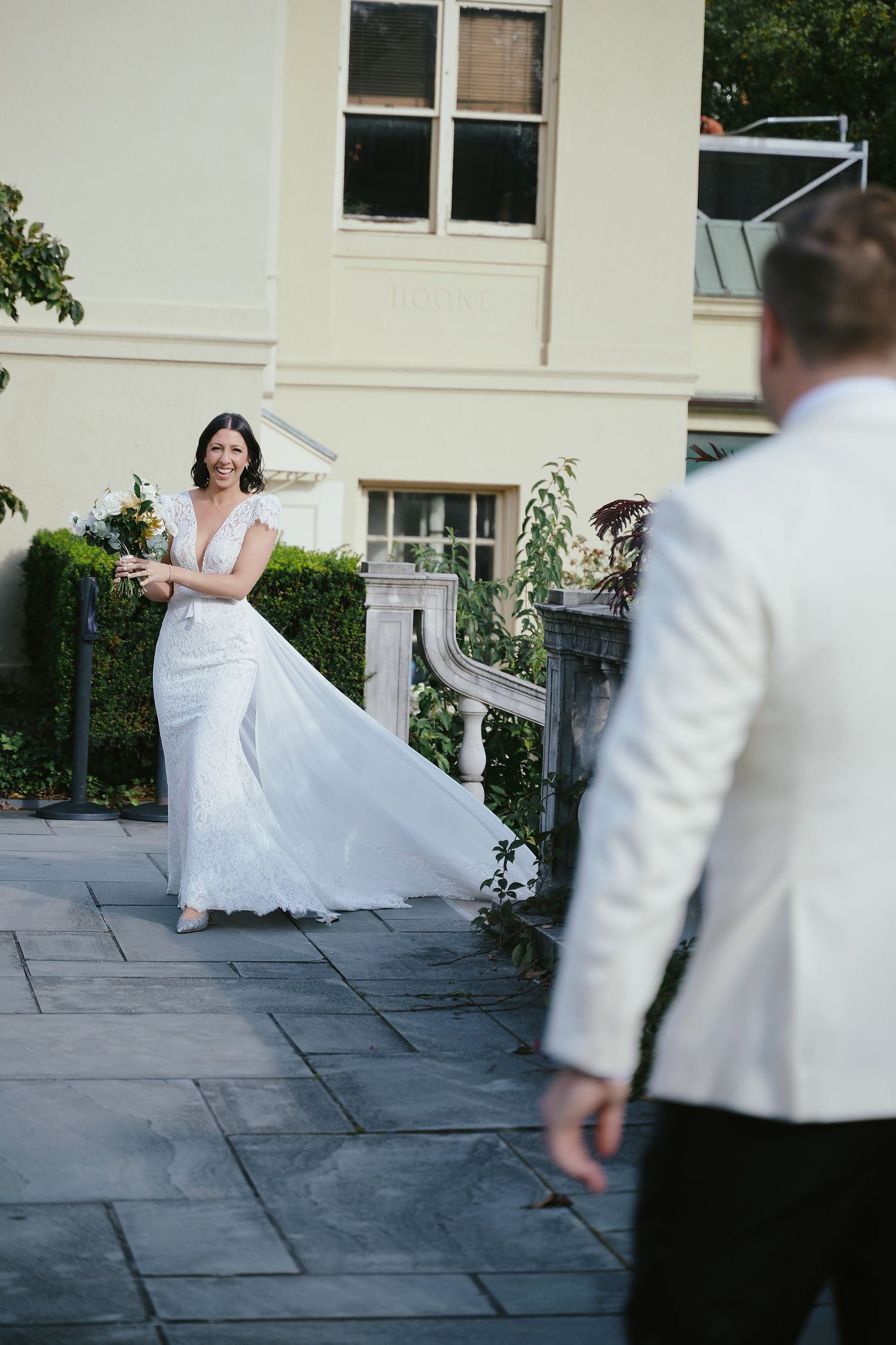 A bride in a flowing white dress twirls on a stone patio while someone in white watches from the side.