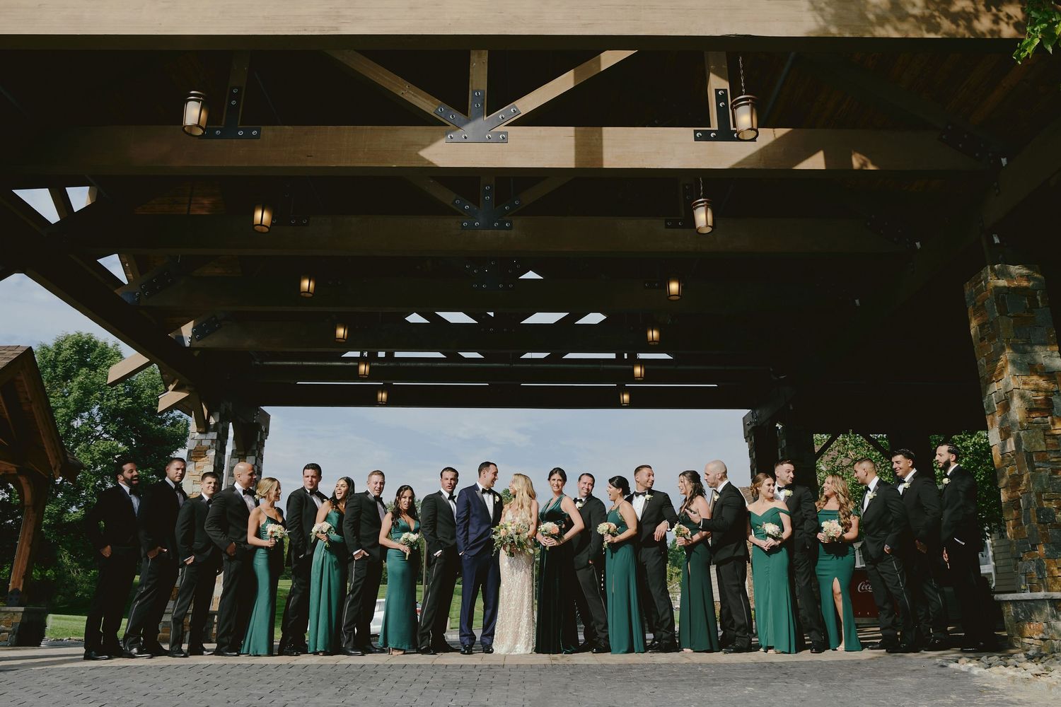 Wedding party stands under rustic wooden pavilion with bridesmaids in matching teal dresses for outdoor ceremony.