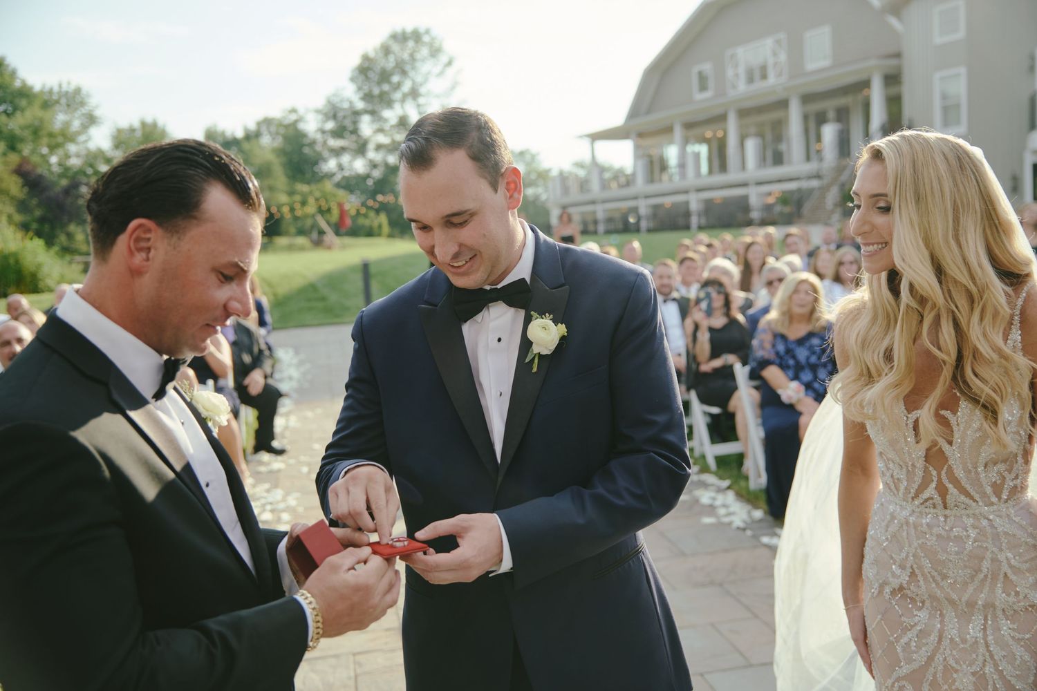 Wedding ceremony guests gather outdoors as rings are exchanged on a sunny day with a large house in the background.