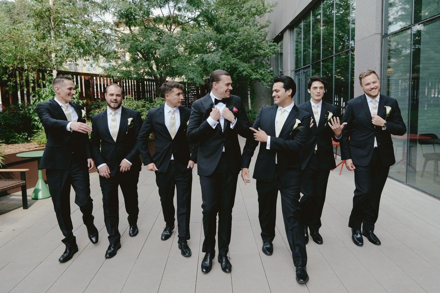 A wedding party in black tuxedos walks and laughs together on a path outside a modern building.