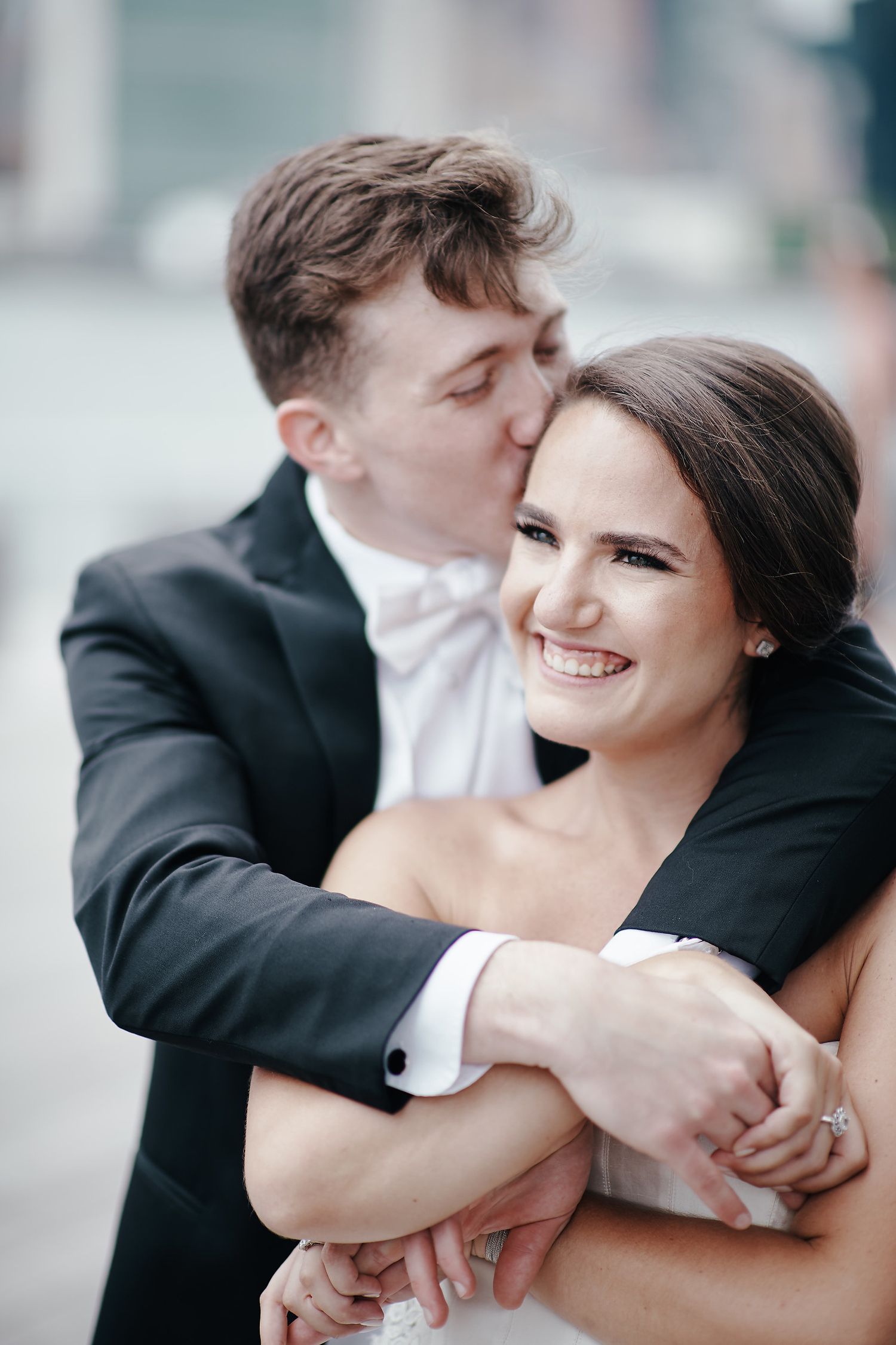 A romantic wedding portrait shows a joyful couple embracing as the groom kisses the bride's cheek outdoors.