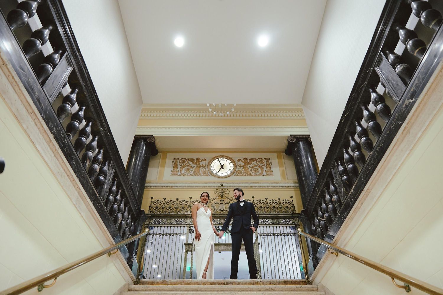A couple stands at the top of an ornate staircase with black railings and recessed lighting in a classic interior setting.
