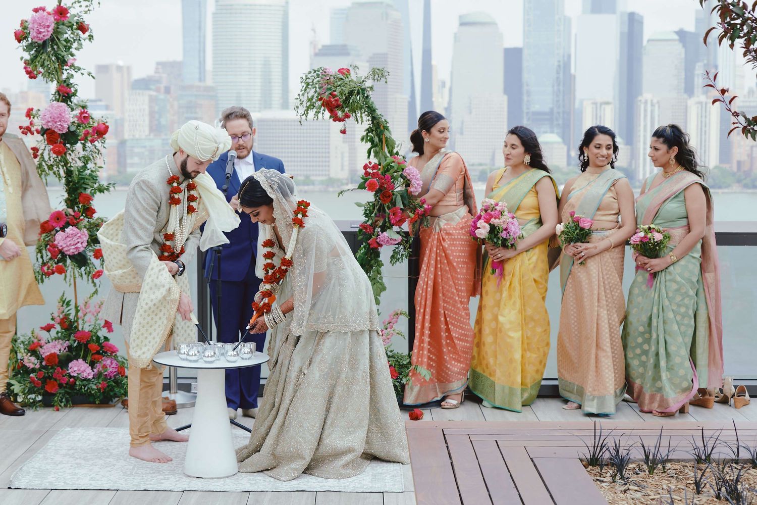 Wedding ceremony with bridesmaids in colorful saris and floral decor on rooftop against city skyline backdrop.
