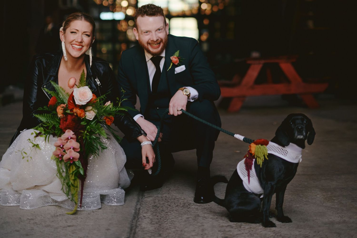 Smiling bride and groom crouching beside their black Labrador dog dressed in a tie at their wedding.