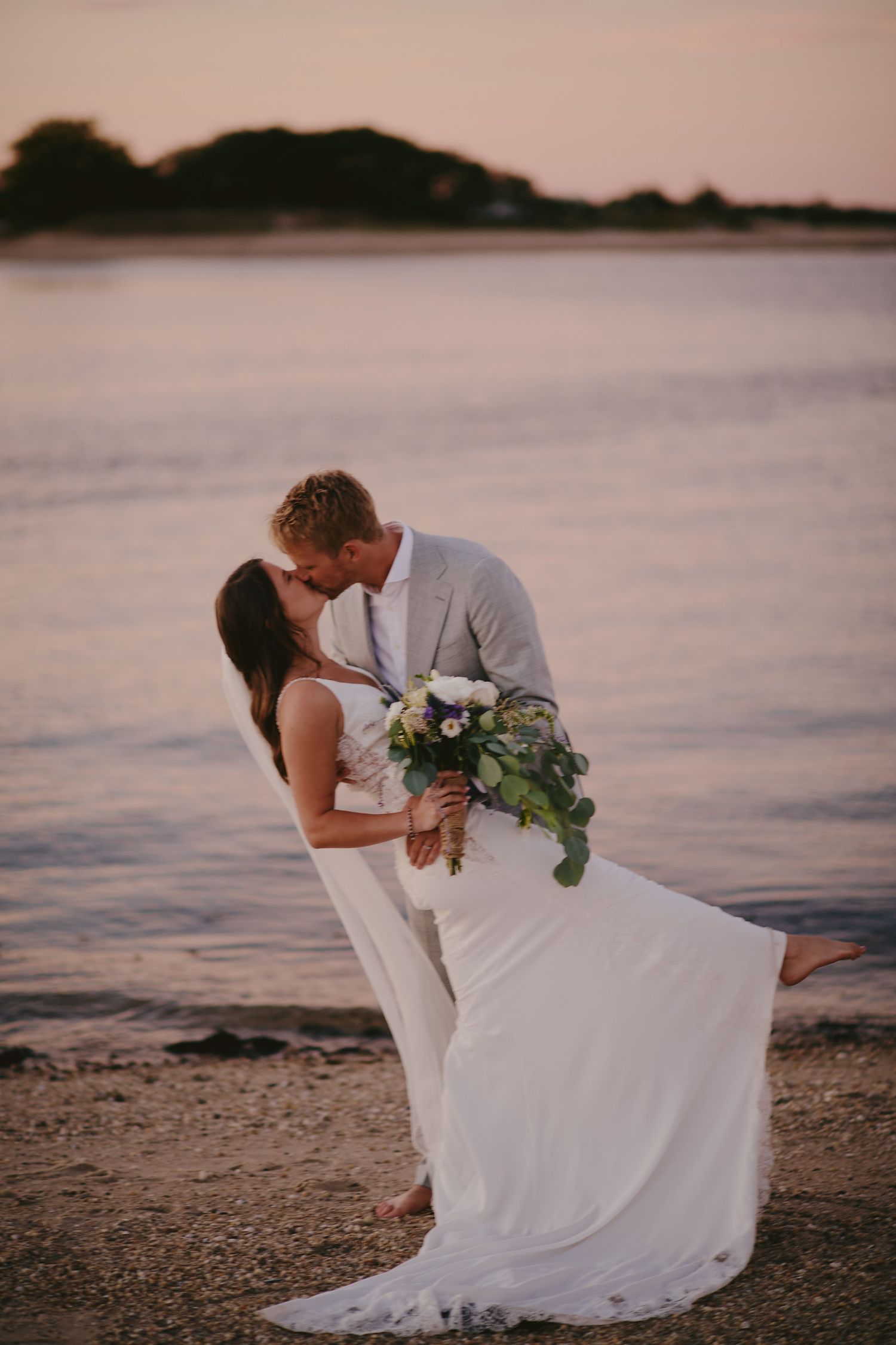 A romantic beachside wedding couple shares an intimate moment at sunset while holding a white and green floral bouquet.