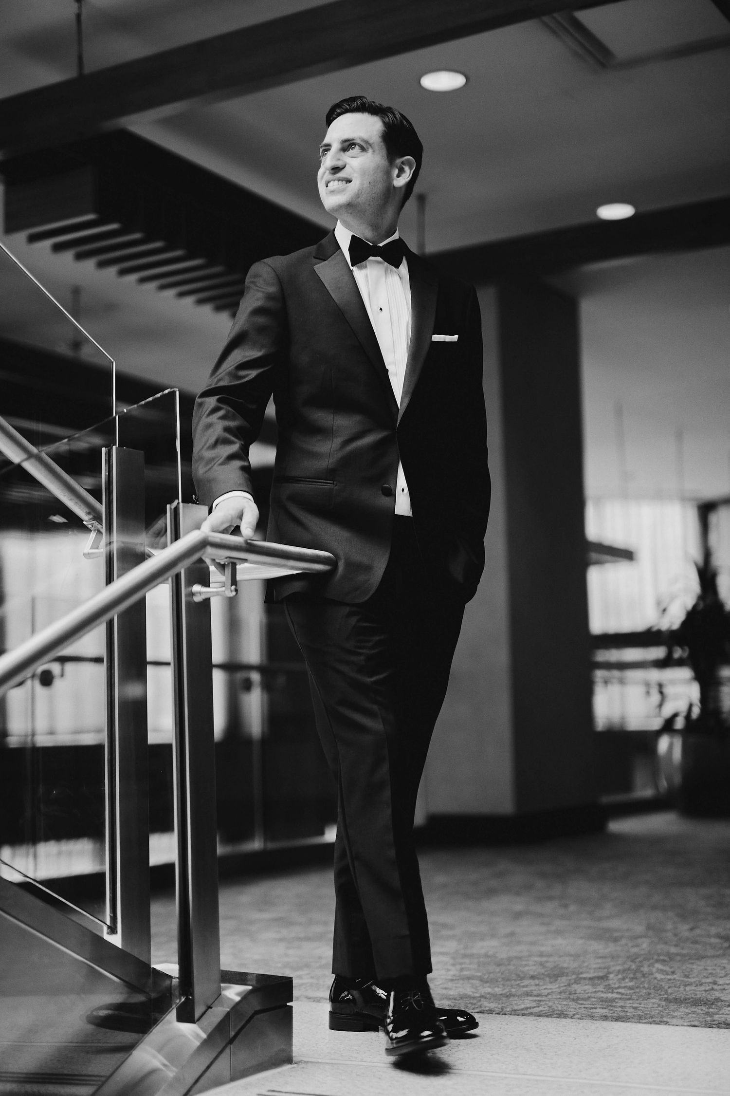 A well-dressed man in a classic black tuxedo and bow tie poses elegantly on a staircase in an indoor setting.