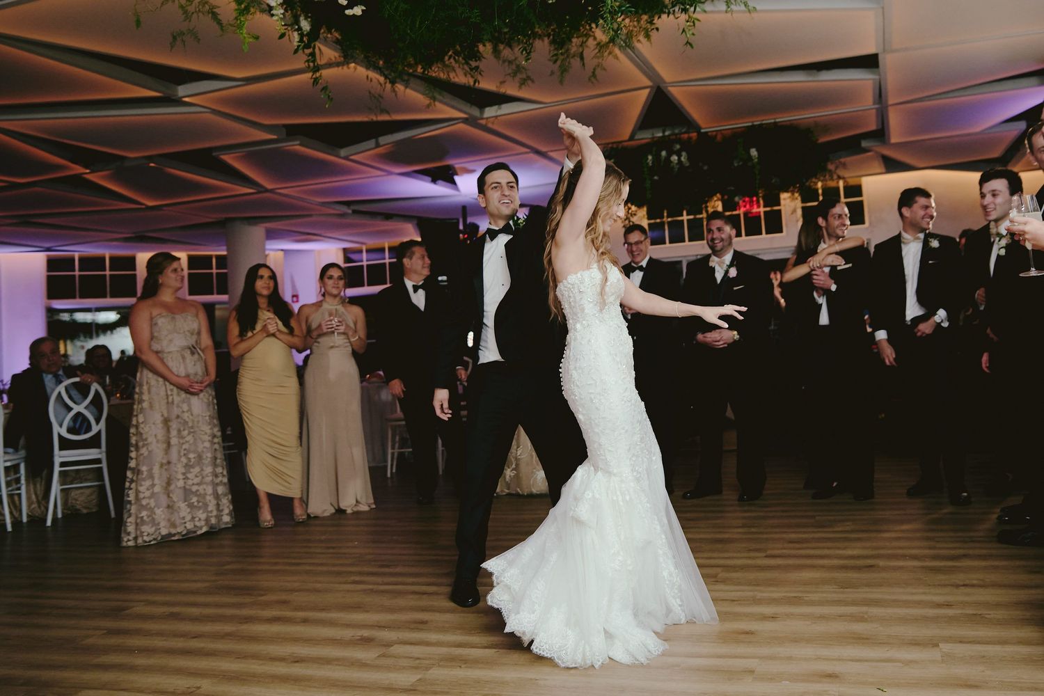 Couple dancing elegantly in a wood-floored venue with decorative lighting and guests watching.