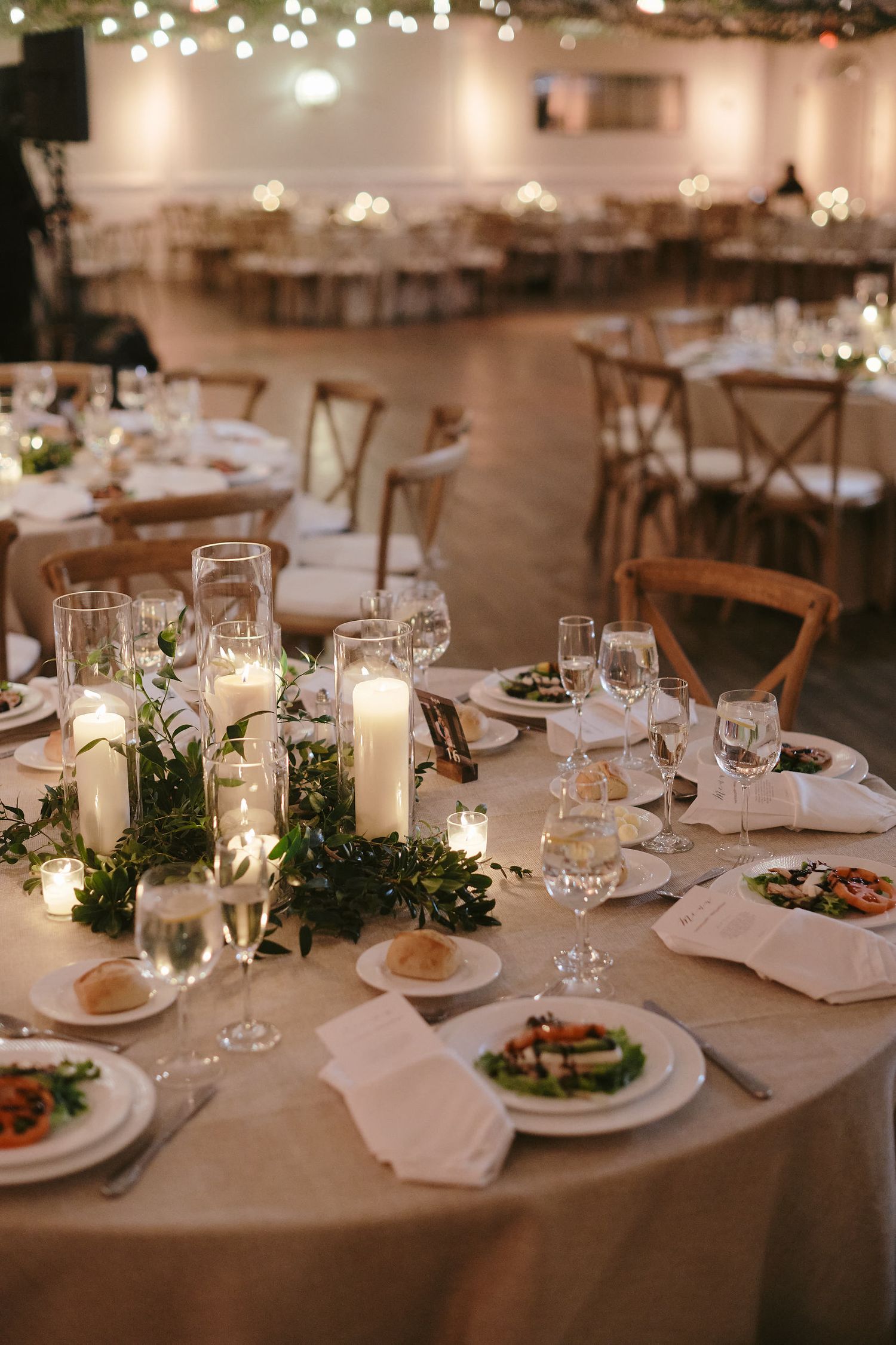 Elegant wedding reception table setting with candles, greenery centerpieces, and place settings on round tables.