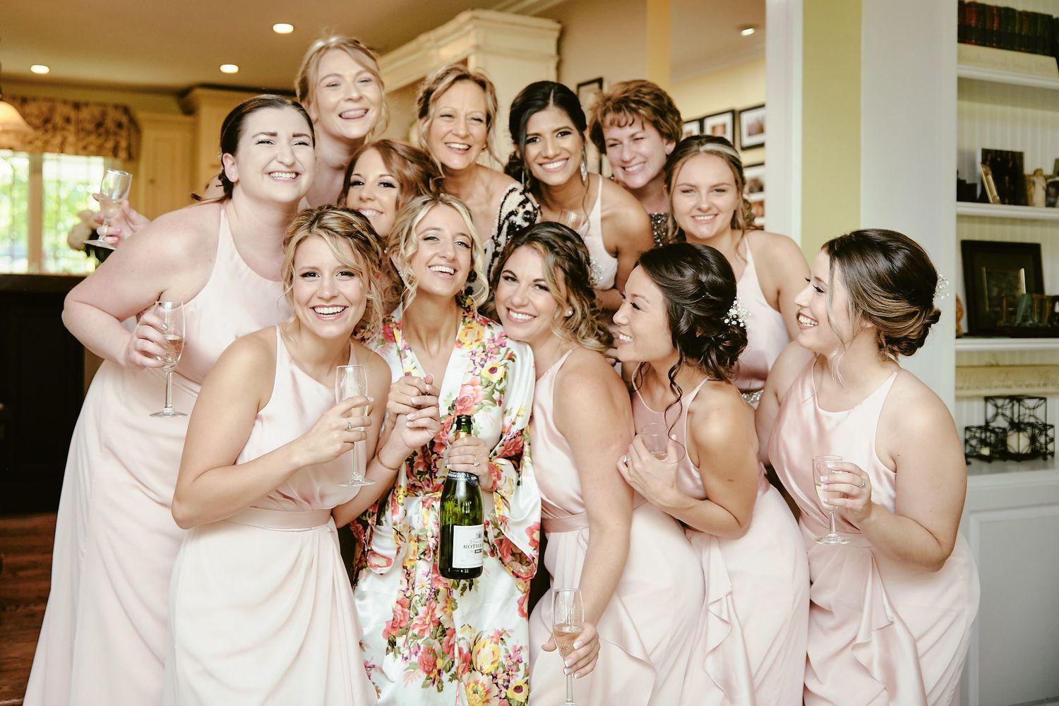 A group wearing blush pink bridesmaid dresses gather around celebrating with champagne at a wedding event.
